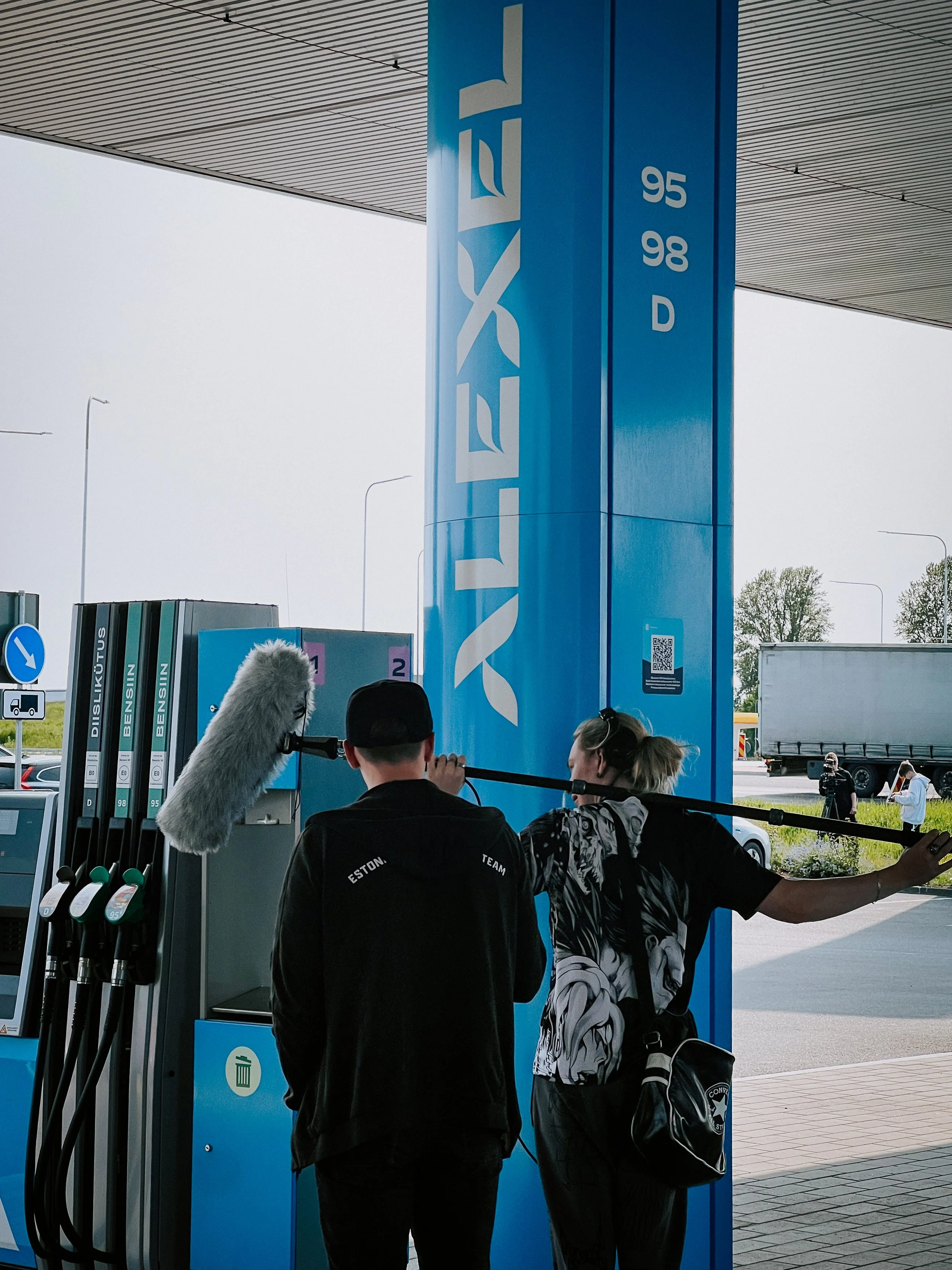 Two people filming a scene at a gas station with a large blue sign that reads "Alexela." One person is holding a boom microphone, and the other is standing nearby with sunglasses on her head and a black bag.