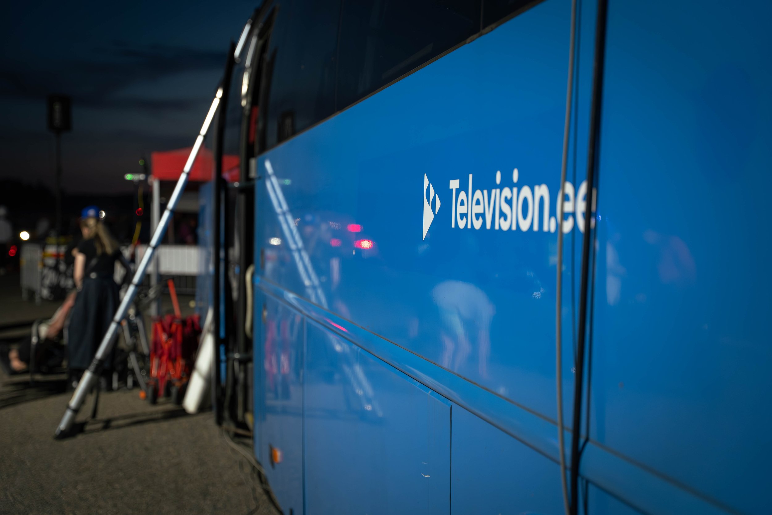 A blue television network bus parked at night with the logo 'Televisioon.ee' visible on its side. In the background, a person wearing a safety helmet is near equipment, possibly on a TV production set.