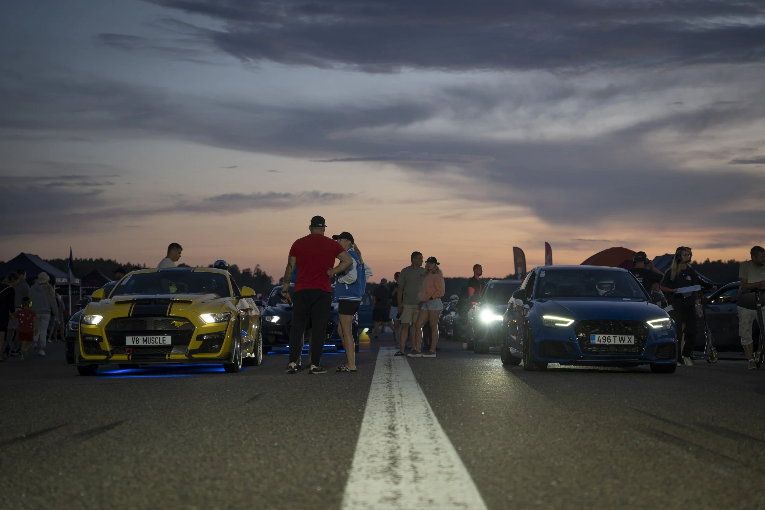 A car meet at dusk, featuring a yellow muscle car on the left and a blue sports car on the right, with people walking and talking around them.