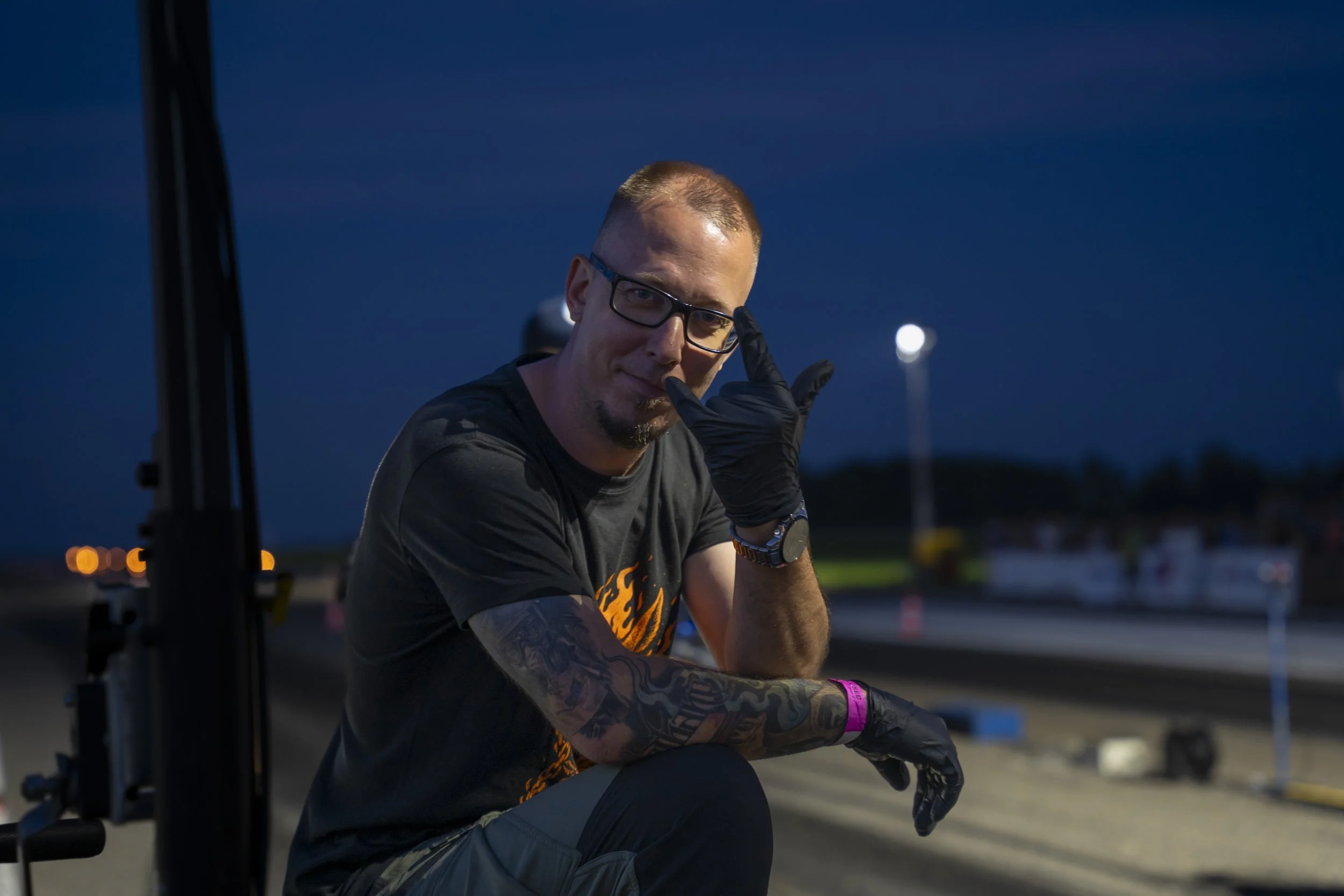 A man with glasses and tattoos kneeling at a racetrack during nighttime, wearing black gloves and making a gesture with his hand near his face.