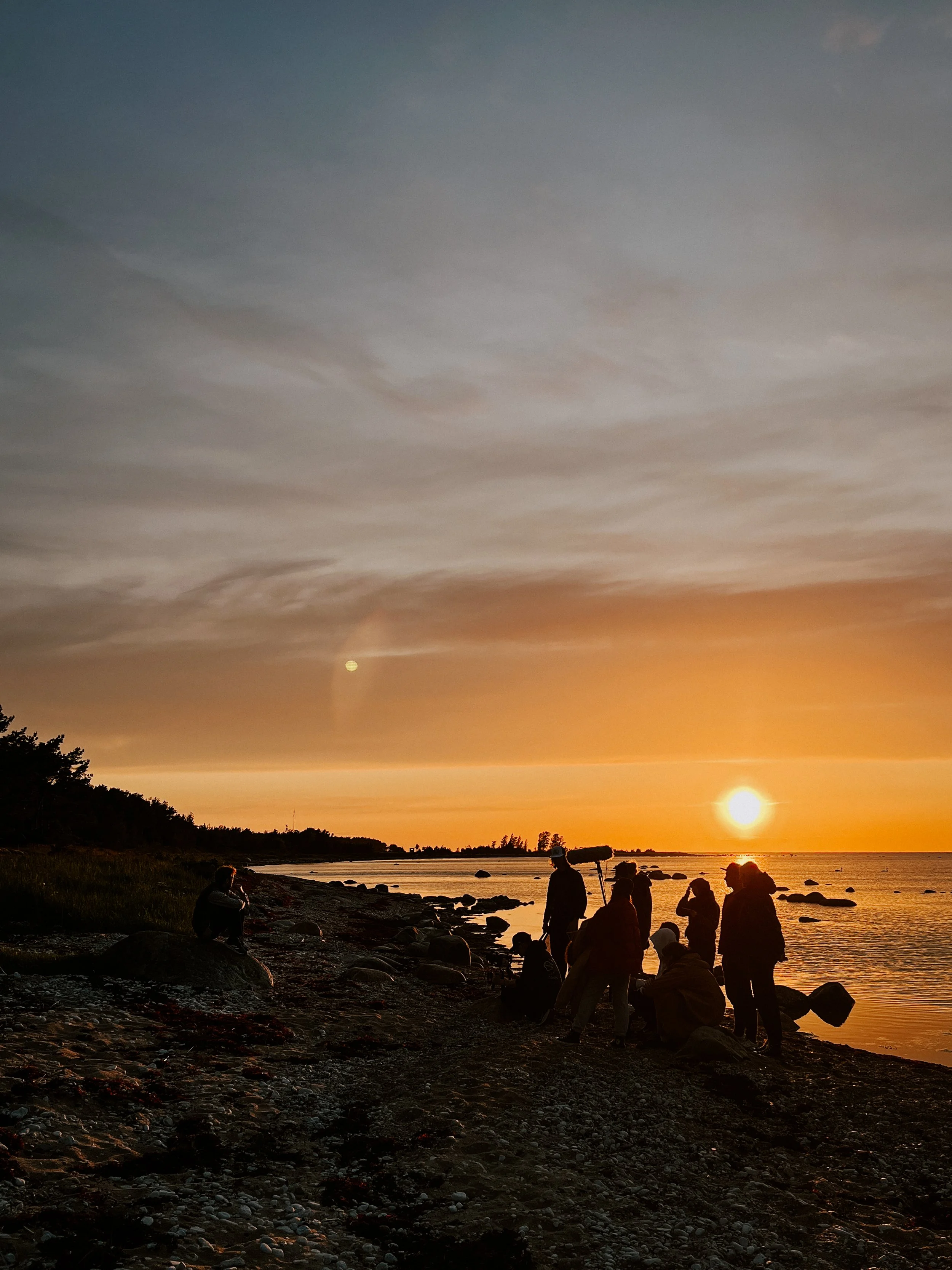 People gather on a rocky beach at sunset, with the sky partially cloudy and the sun near the horizon, some taking photos, others sitting or standing with camera equipment.