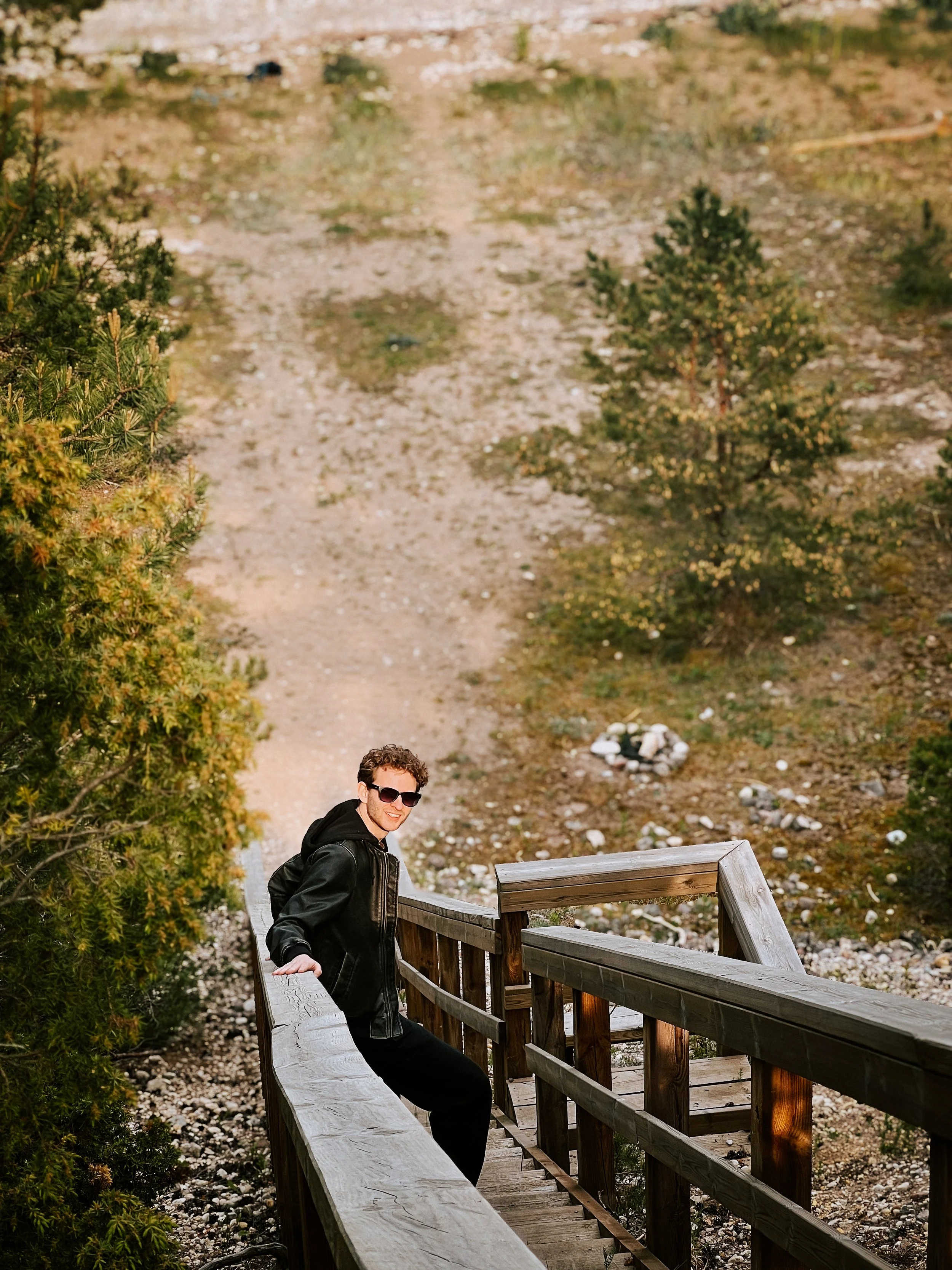 A young man with curly hair, wearing sunglasses, a black leather jacket, and black pants, sitting on a wooden railing of a staircase outdoors, smiling at the camera with trees and a dirt path in the background.