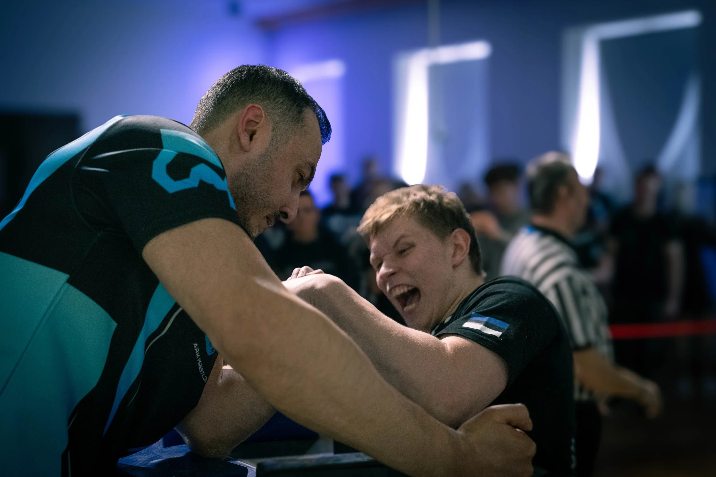 Two young men are engaged in an arm wrestling match at a competition or event, with one appearing determined and the other smiling. Several people are watching in the background.