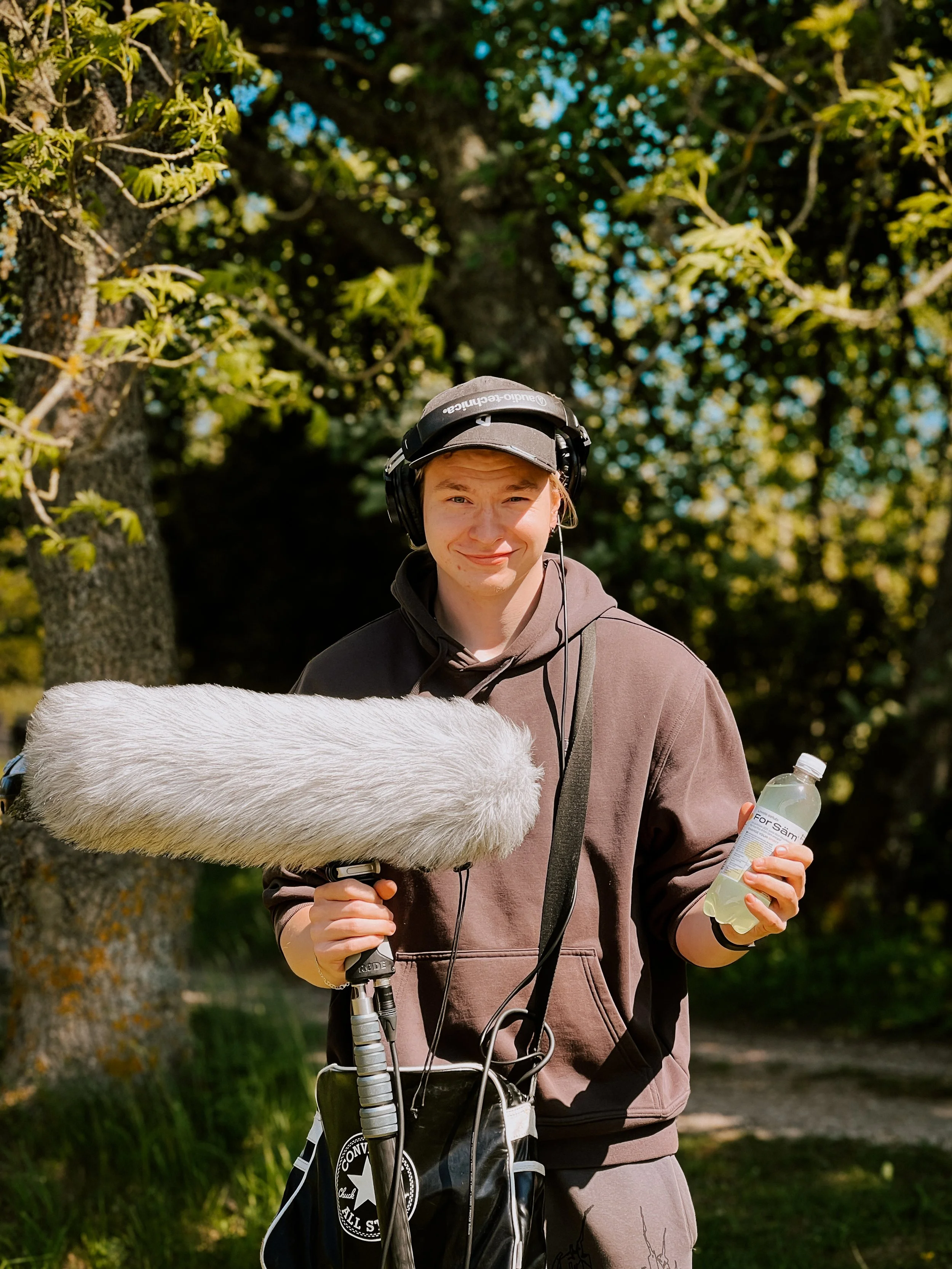Young man with headphones holding a boom microphone and a water bottle outdoors surrounded by trees.