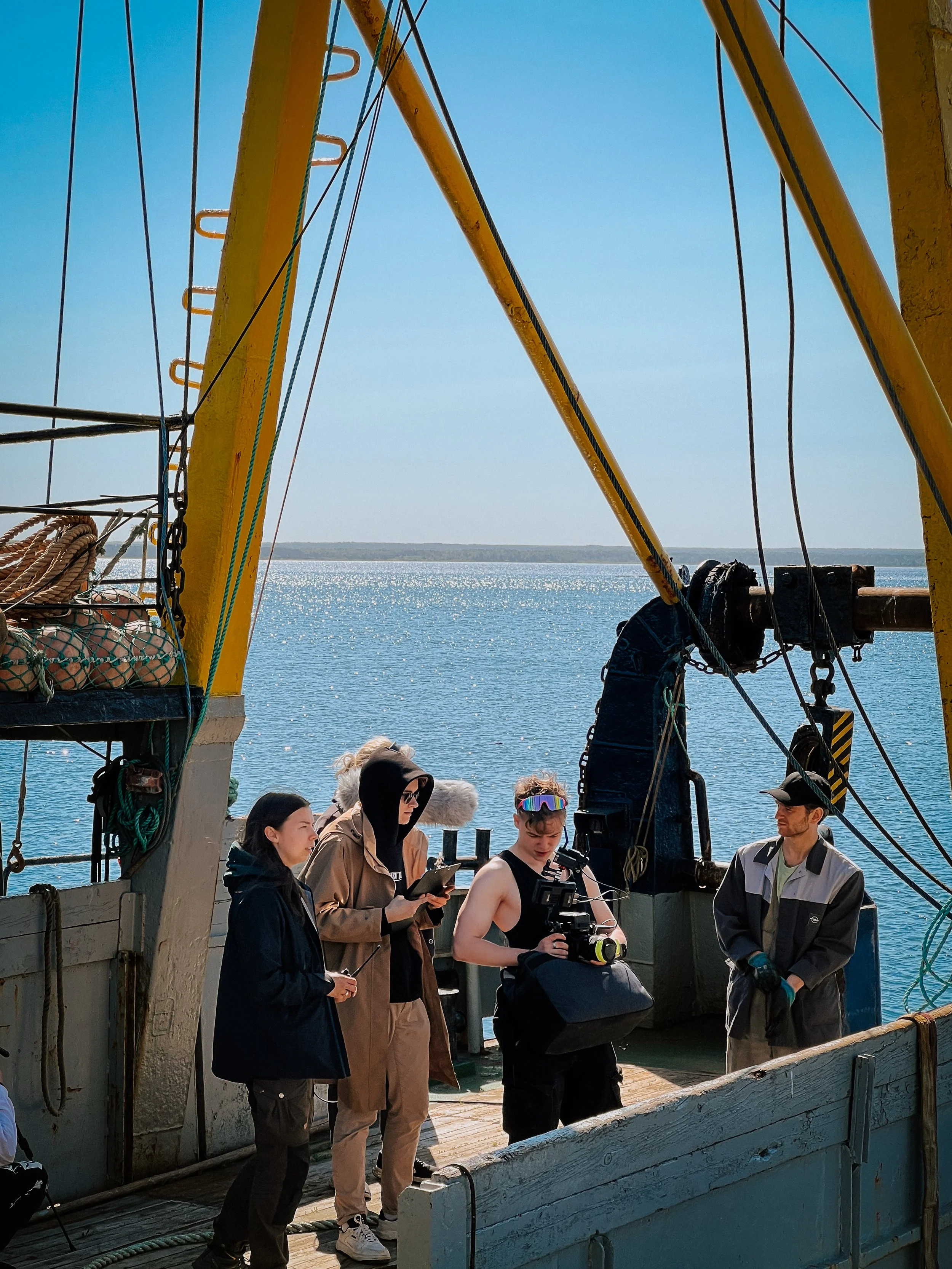 A film crew preparing to shoot on a boat by the water on a sunny day, with four people, including a camera operator and director, gathered on the deck.