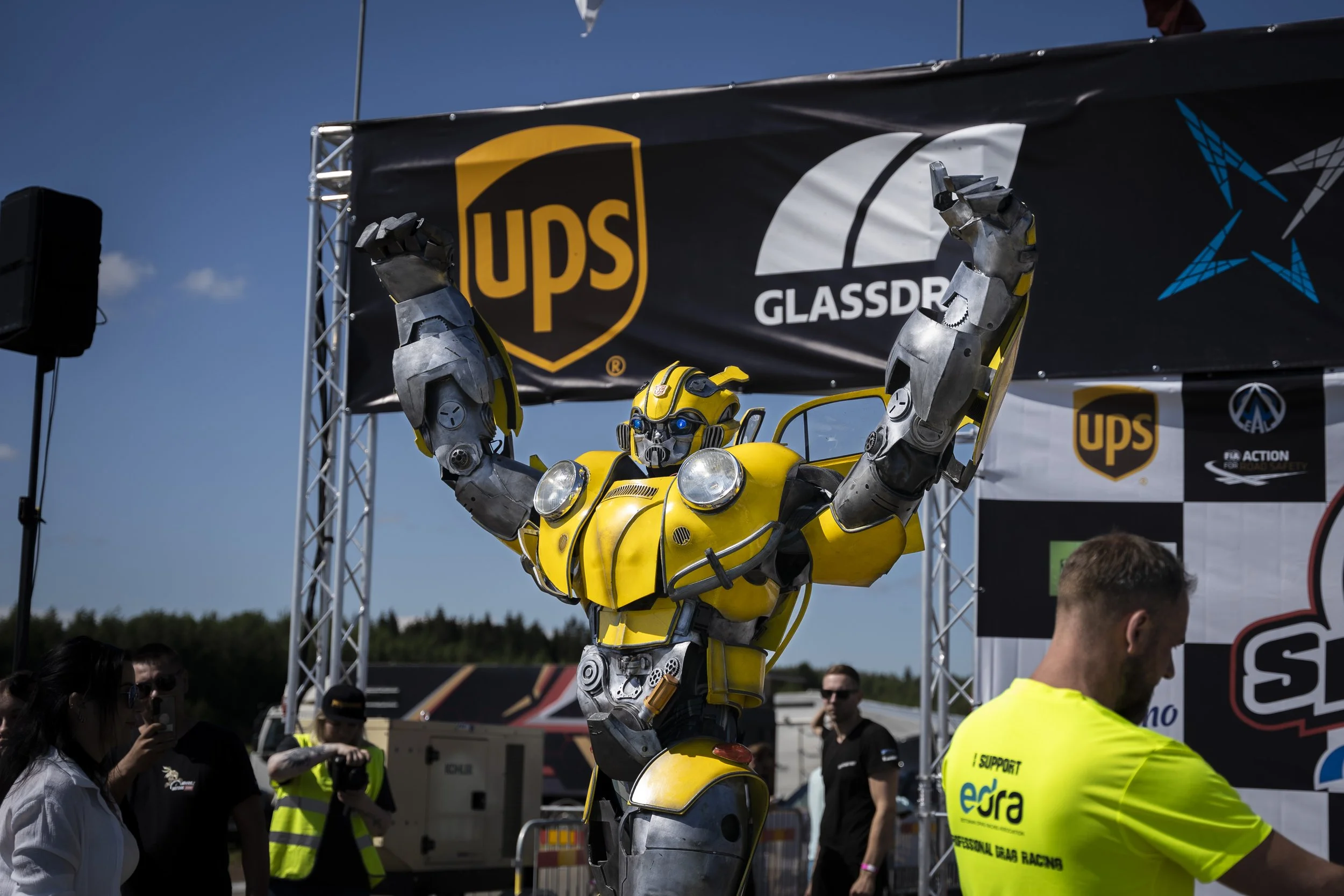 A person dressed as Bumblebee, a Transformer character, with yellow and gray robotic costume, raising their arms in front of a stage with sponsor banners including UPS and Glassdoor, at an outdoor event on a sunny day.