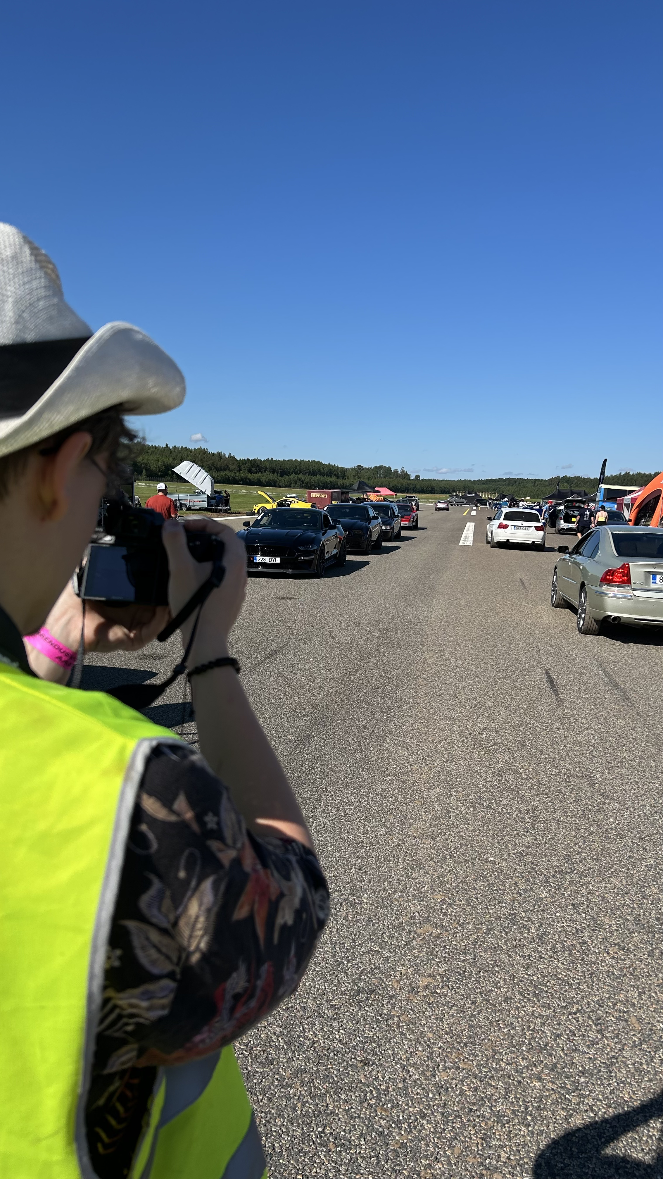 Person in a yellow vest taking a photo of a lineup of cars at a racetrack on a sunny day.