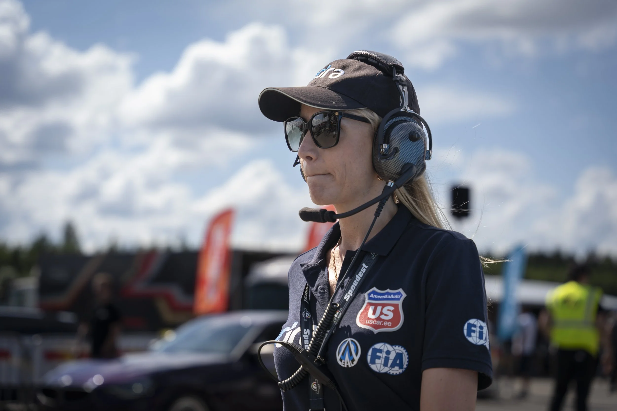 A woman wearing sunglasses, a black cap, and a headset with a microphone at a racing event with cars and flags in the background.