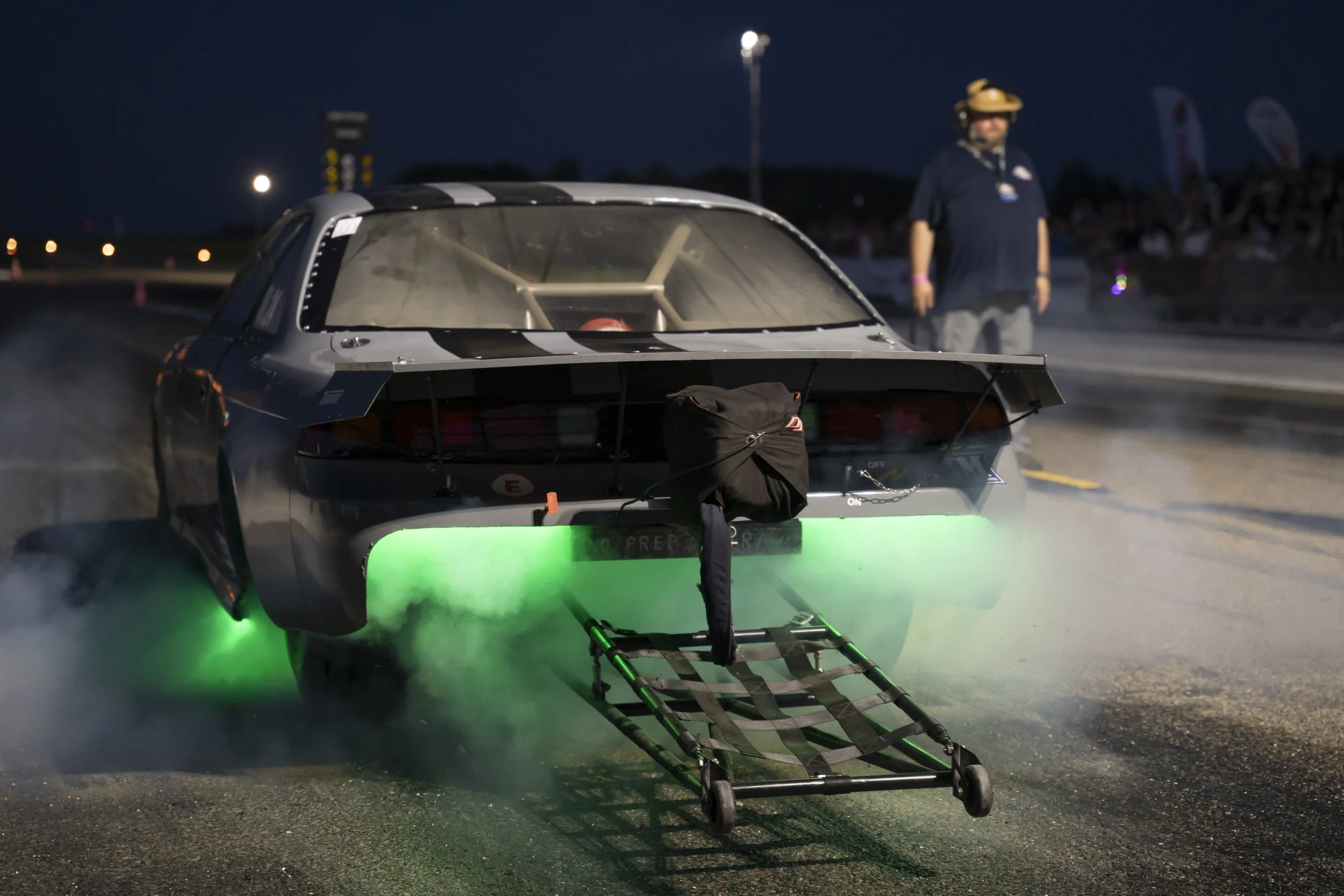 Nighttime drag racing car with green underglow lights and a wheel chair ramp, with a person in a helmet standing next to it.