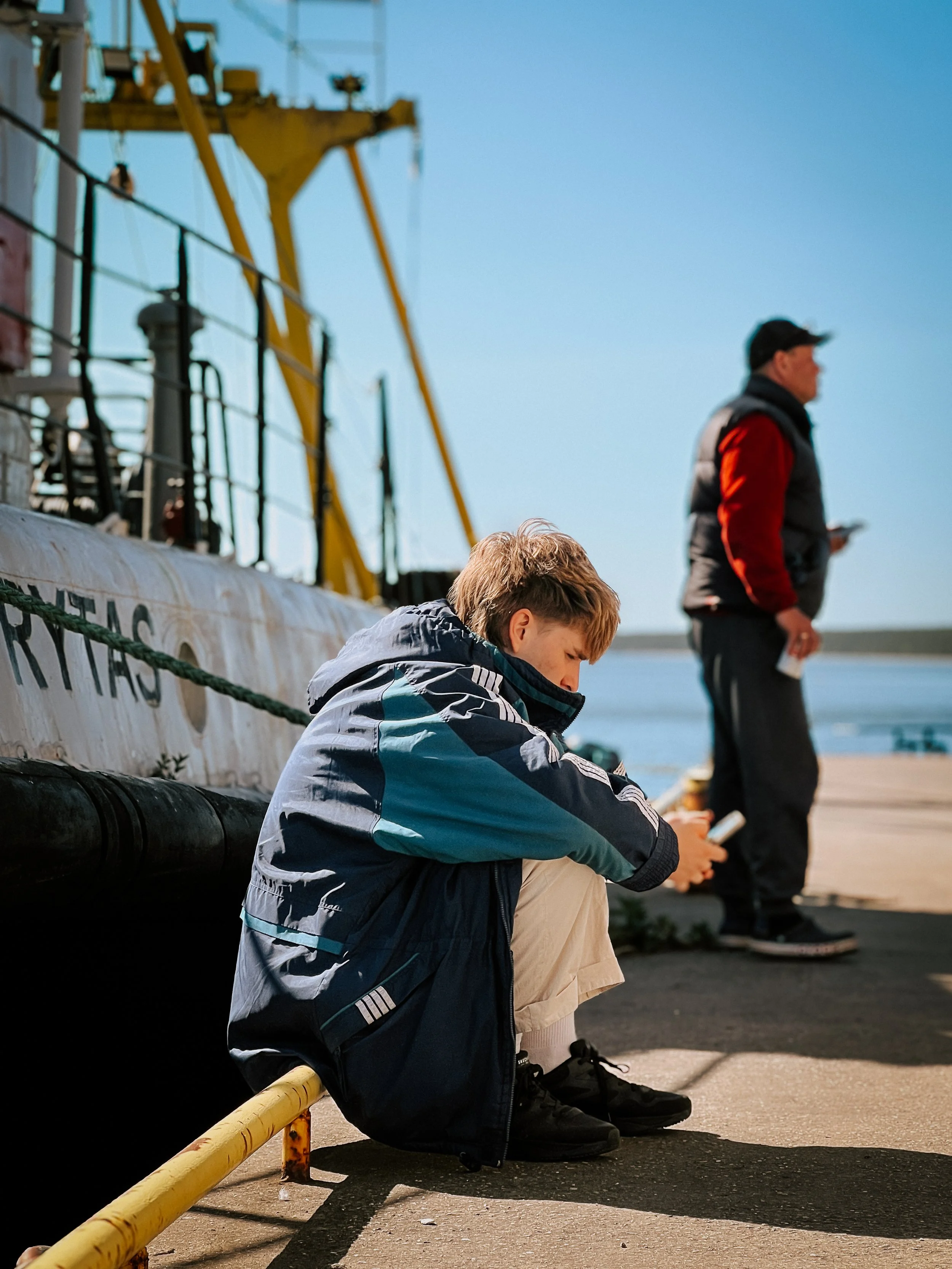A young boy is sitting on the ground near a dock, focused on his phone. In the background, an adult man stands, also near the dock, looking at his phone. The setting appears to be at a harbor or marina with a large boat and yellow crane in the backgr