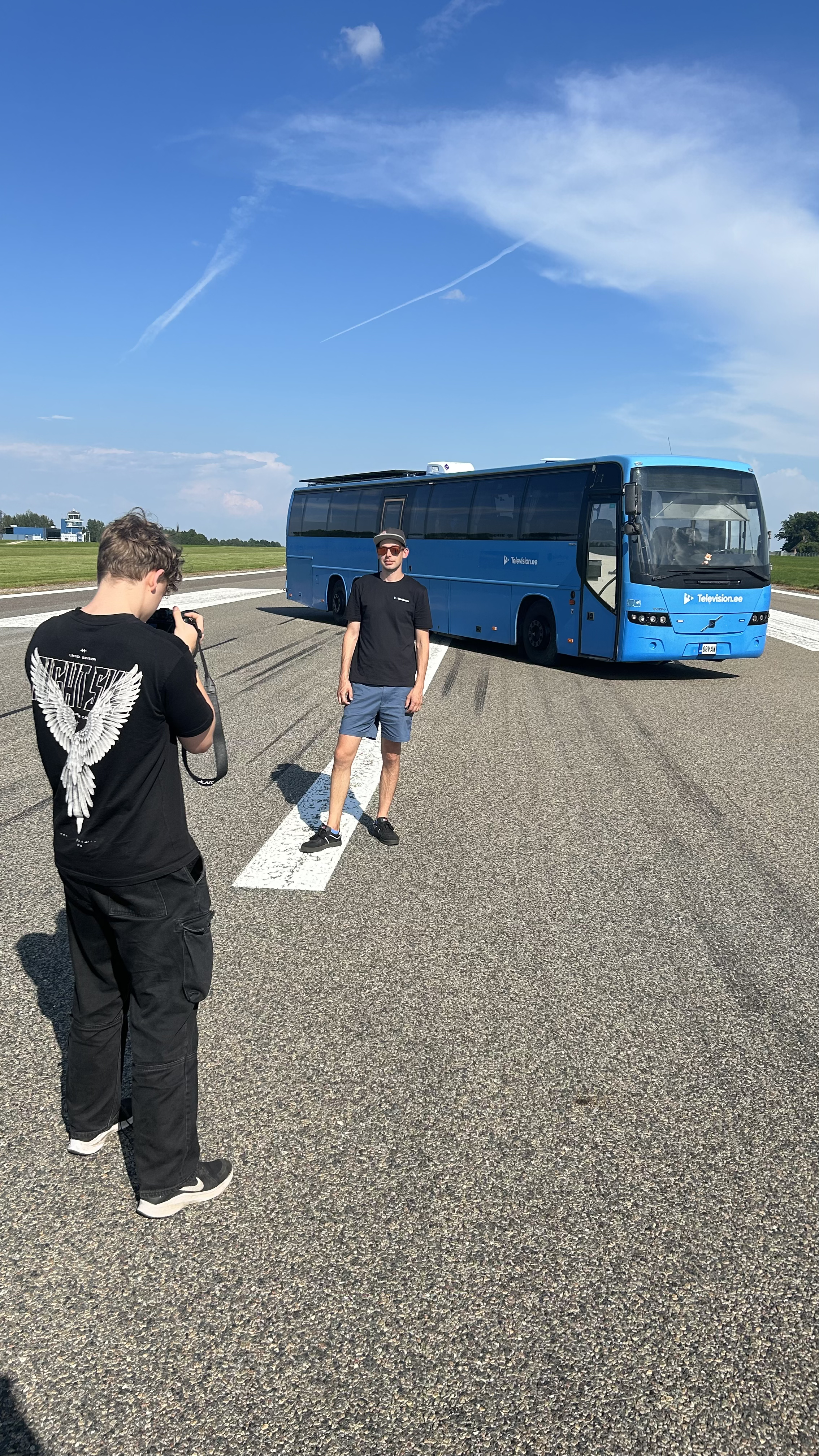 A young man stands in front of a blue bus for a photoshoot while another person takes his picture on a bright, sunny day with a clear blue sky.