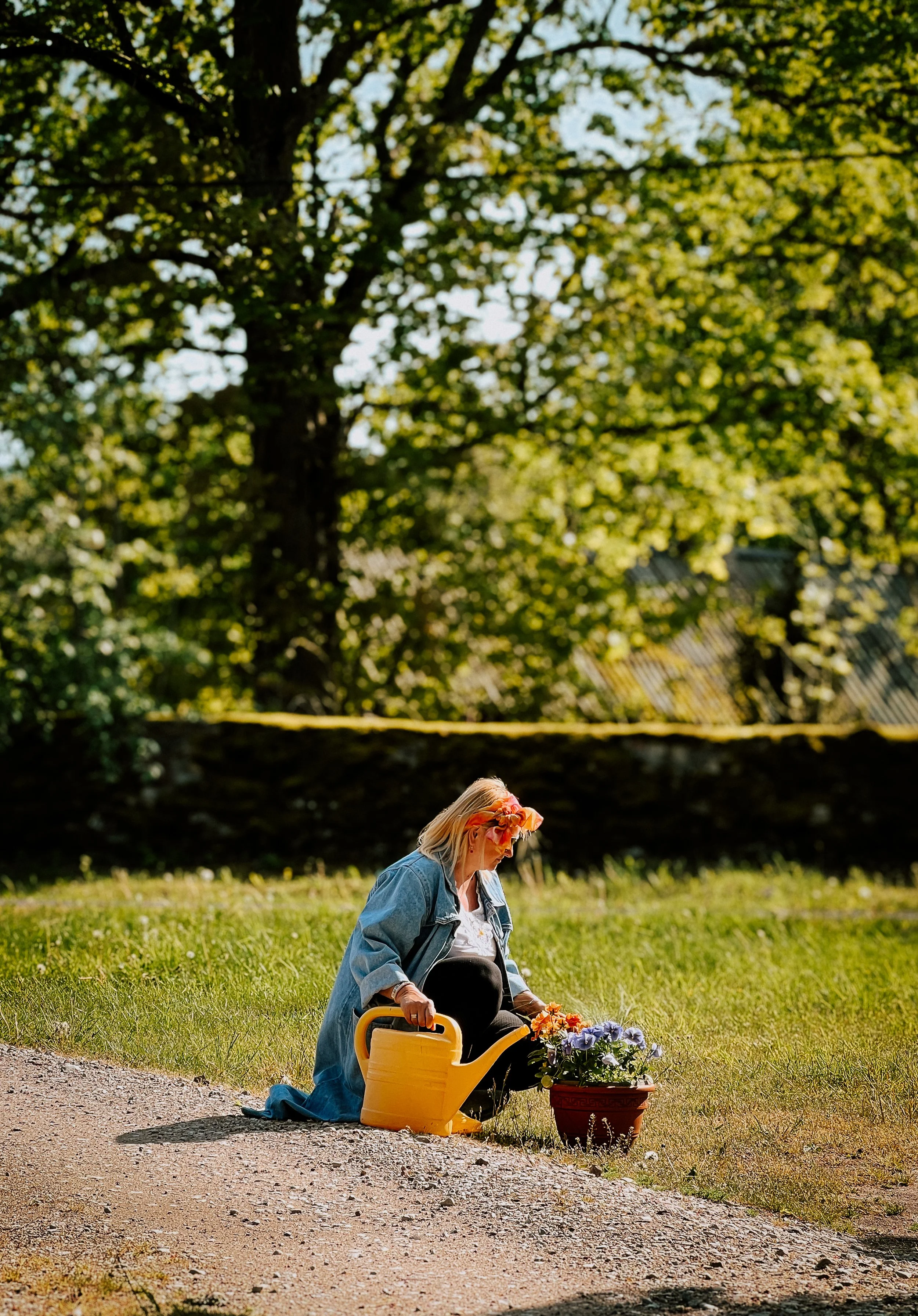 A woman with blond hair, wearing a pink headband, blue oversized denim jacket, and sunglasses, is sitting on the ground next to a yellow watering can and a pot of colorful flowers, outdoors on a grassy area with a large trees and a fence in the backg