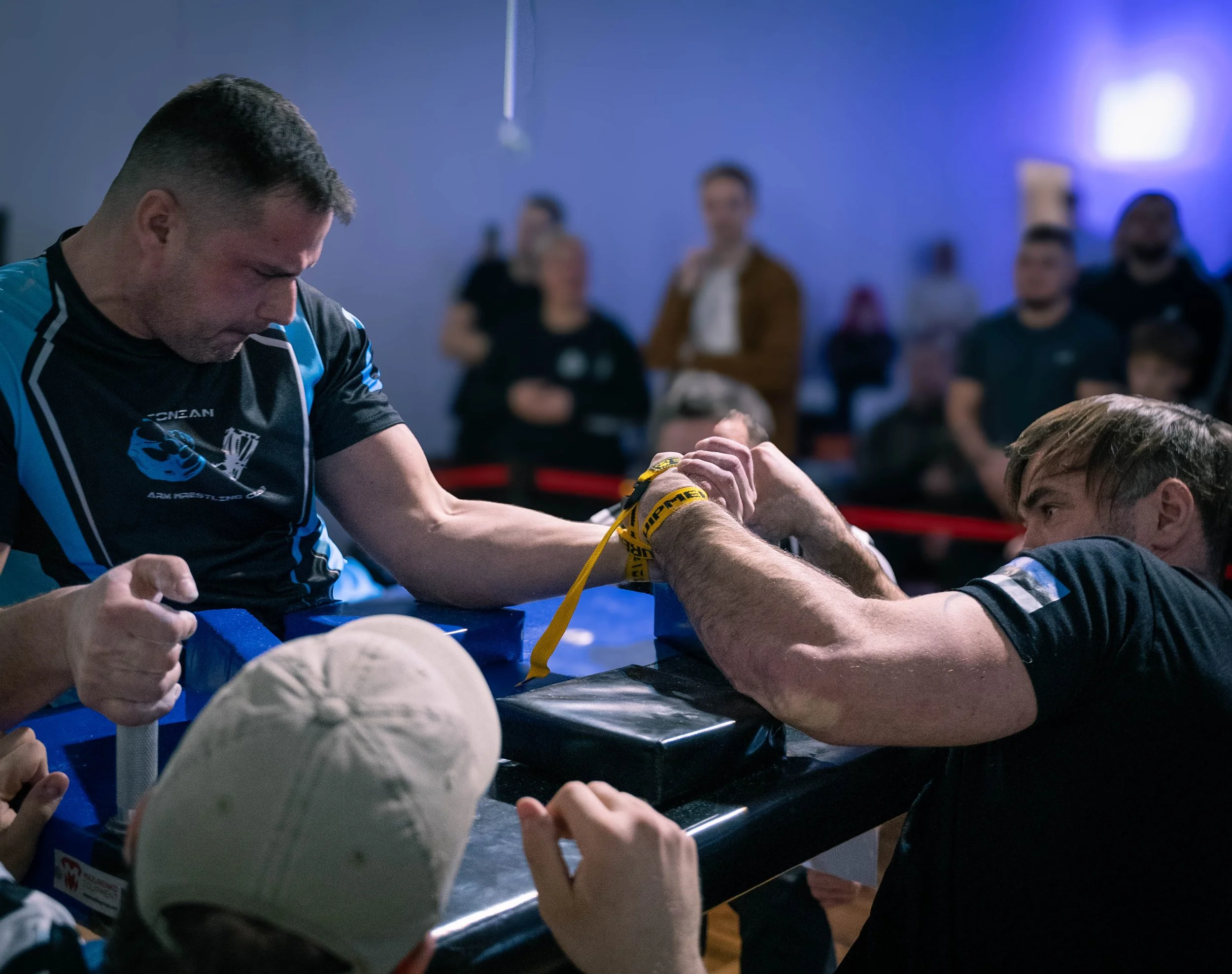 Two men competing in an arm wrestling match while other spectators watch in the background.