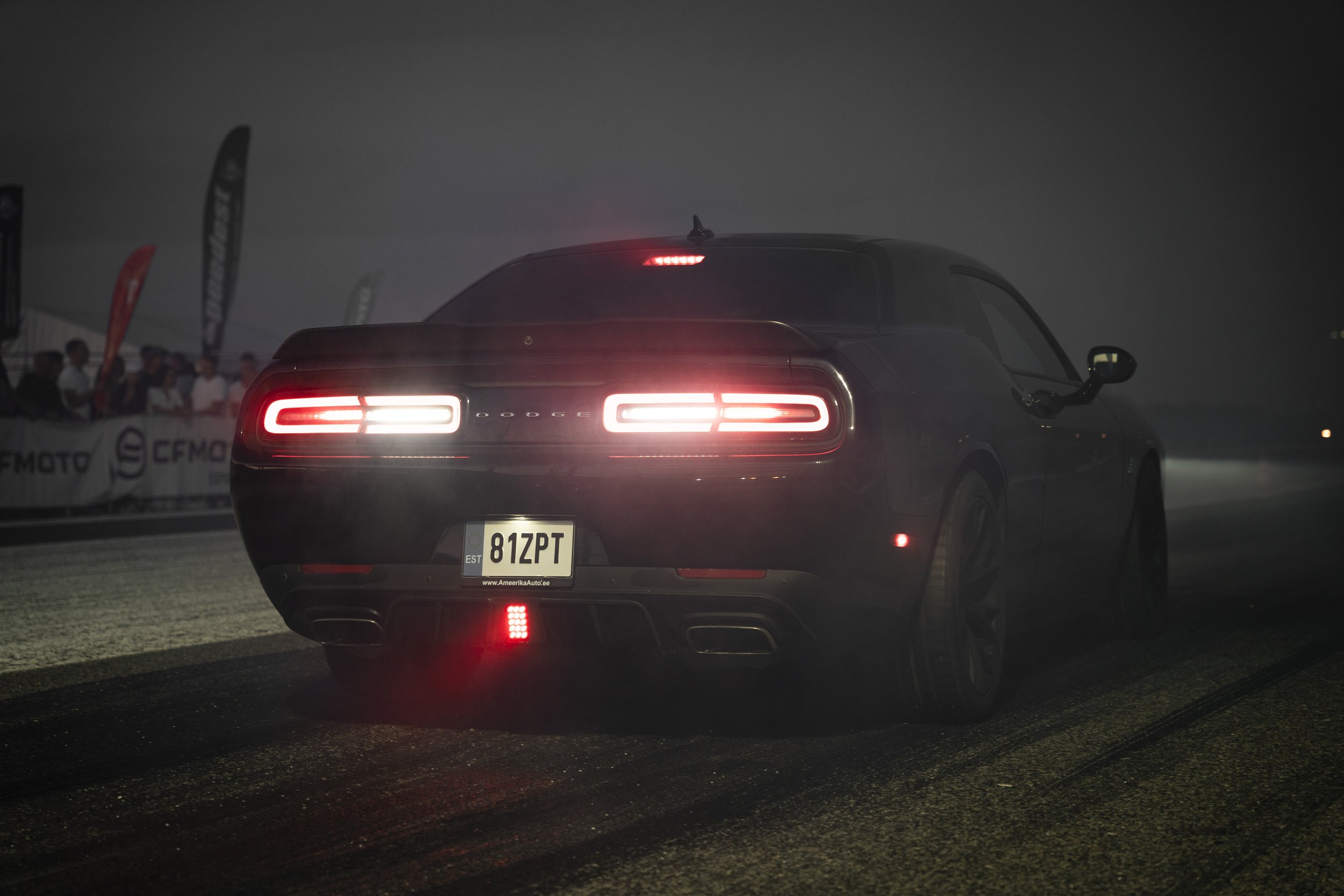 A black Dodge Challenger car at night with glowing red tail lights, license plate 81ZPT, parked on a dark street with a group of people and flags in the background.