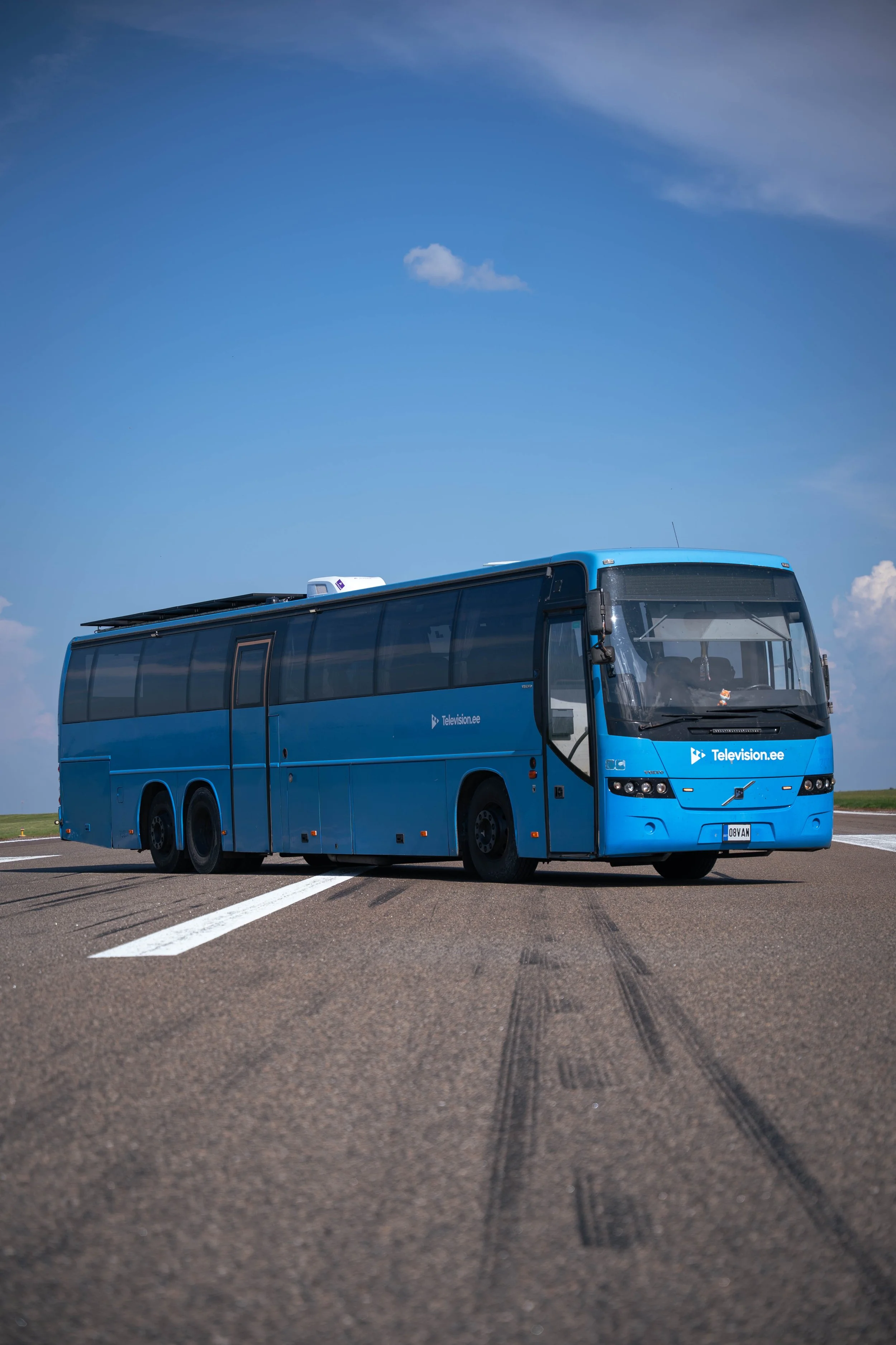 Blue television broadcast bus parked on an open road with tire marks visible on the pavement and a partly cloudy sky in the background.