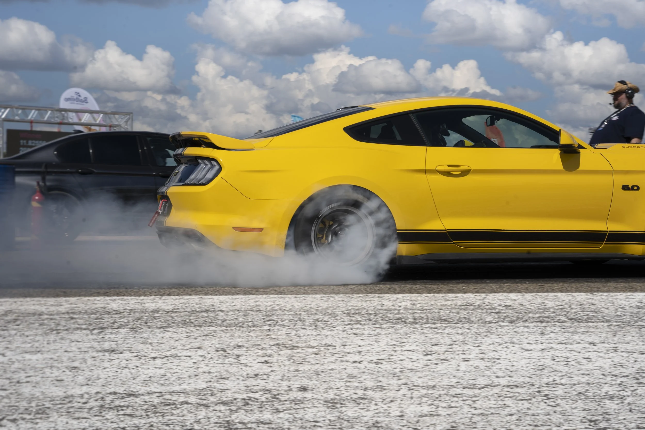 Yellow sports car performing a burnout at a racing event, with smoke billowing from the tires and a black car in the background, against a partly cloudy sky.