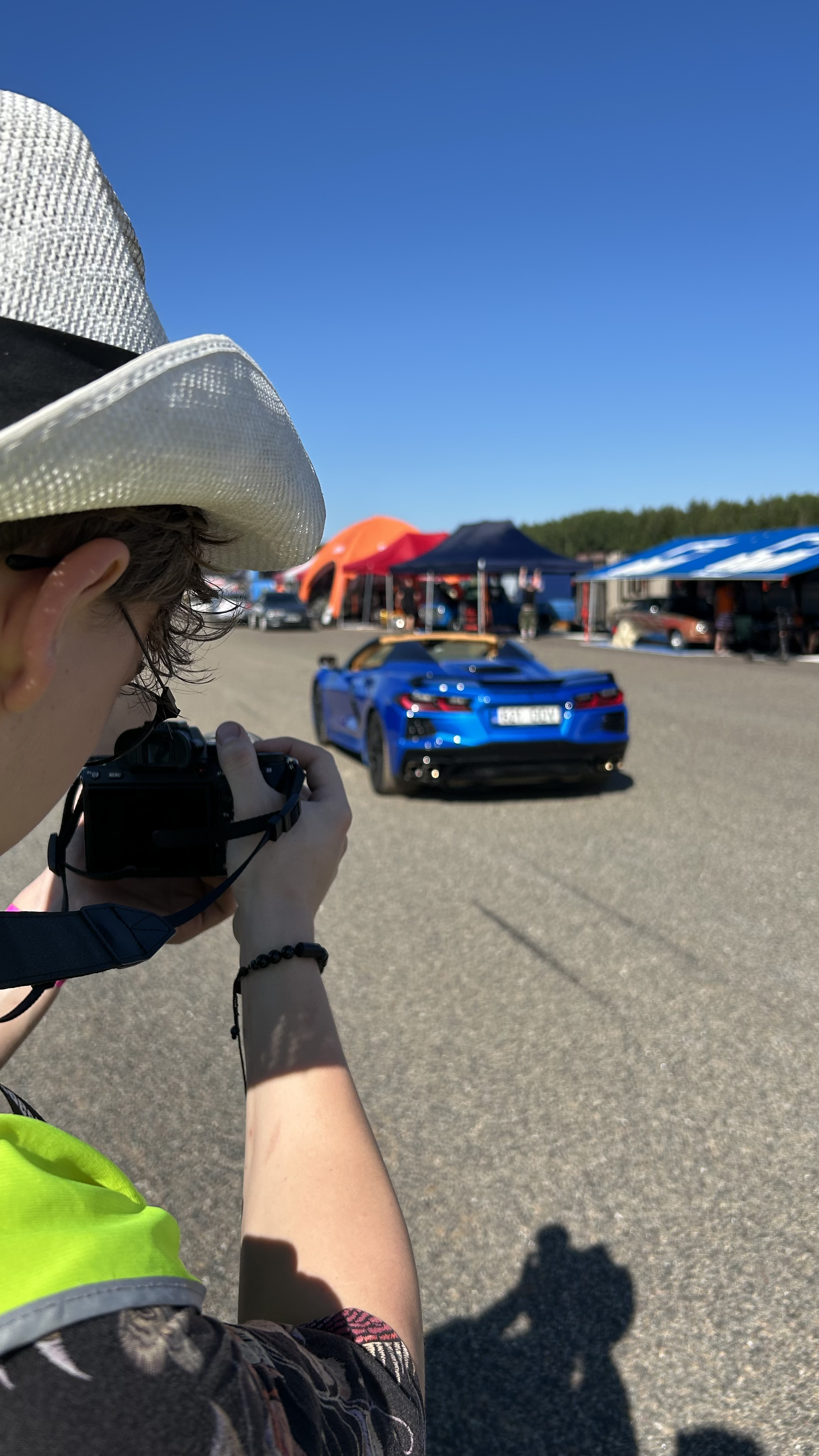 A person taking a photo of a blue sports car with a convertible top at an outdoor event on a sunny day, with tents and cars in the background.