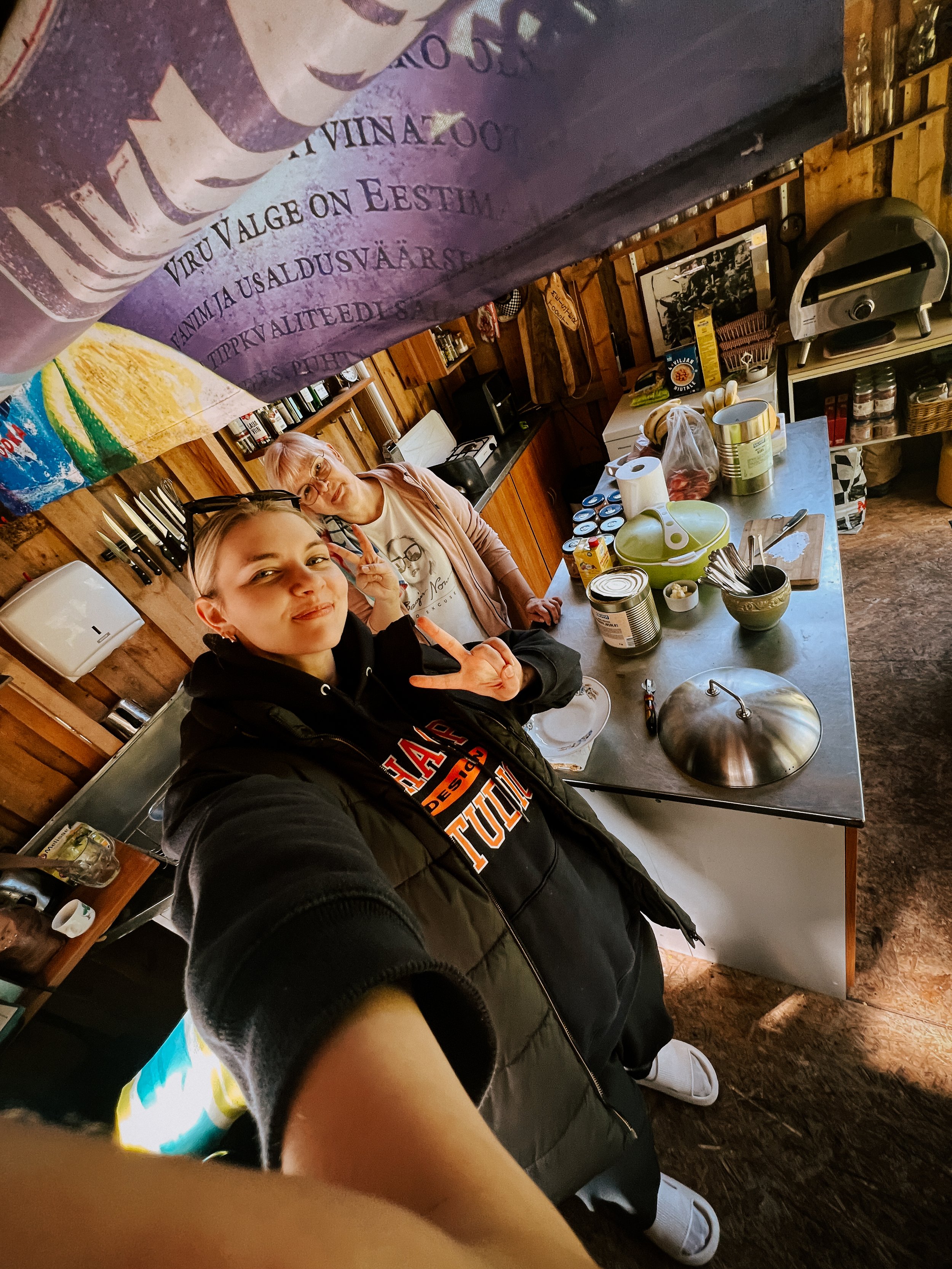 Two women taking a selfie in a rustic kitchen with wooden walls. One woman is in the foreground making a peace sign, and the other woman is in the background also making a peace sign. The kitchen has various food items and kitchen utensils on the cou
