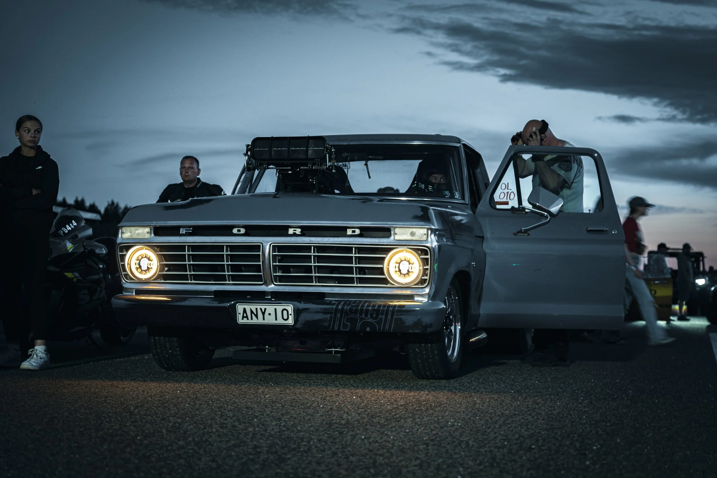 A vintage Ford race car with its headlights on, parked on a dark road during dusk, with people standing around and a dramatic cloudy sky overhead.