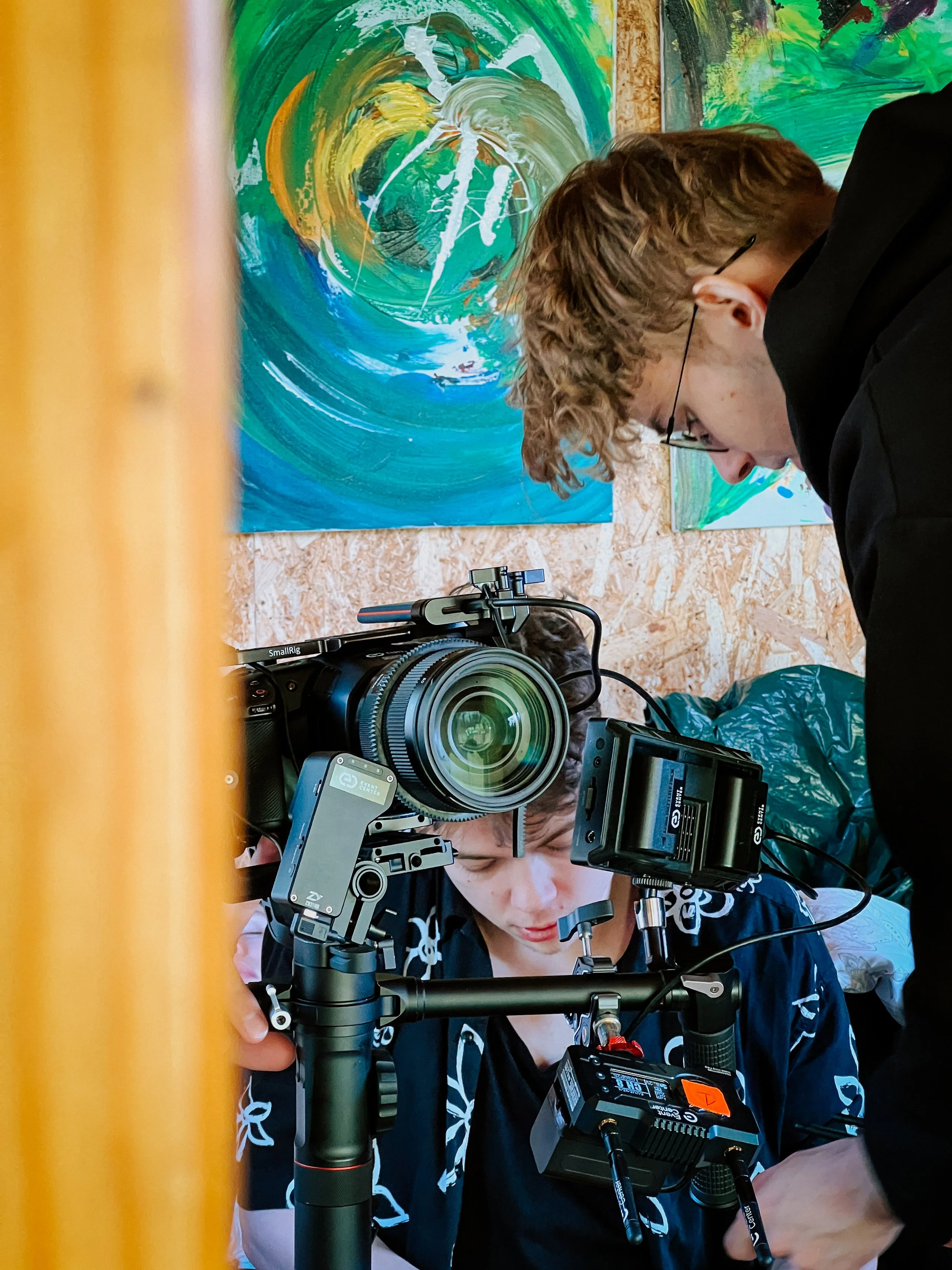 Two people setting up filming equipment around a person, with colorful abstract paintings on the wall in the background.