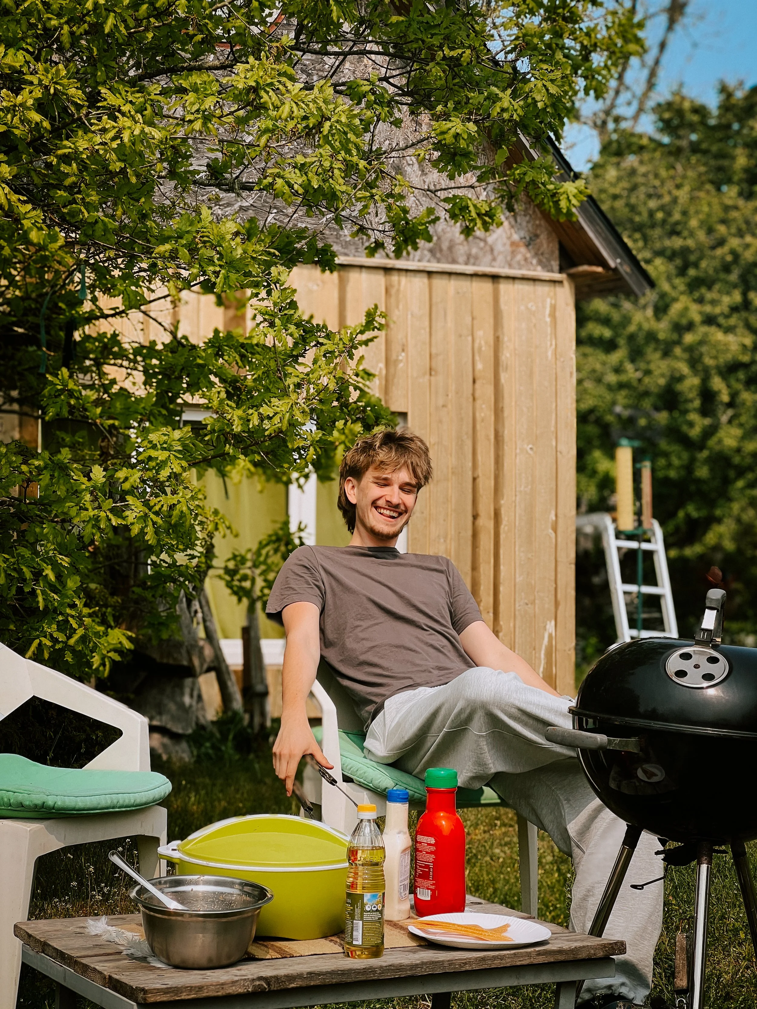 A young man with brown hair smiling and sitting relaxed in a white outdoor chair near a grill surrounded by condiments, utensils, and outdoor furniture, with trees and a wooden shed in the background on a sunny day.