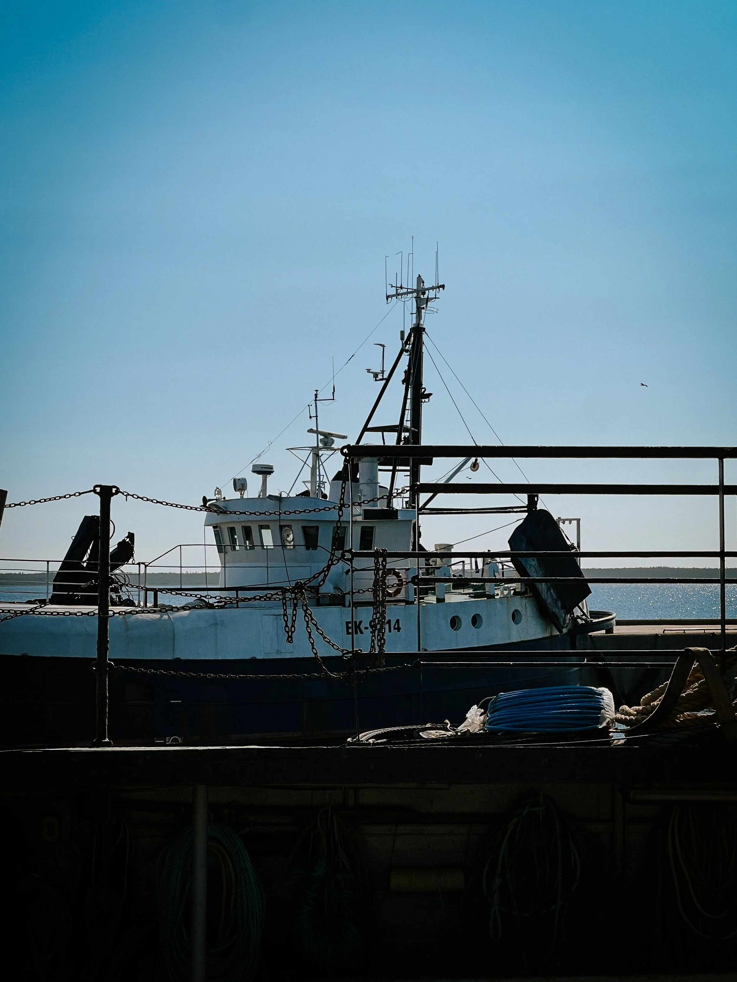 A white and blue fishing boat docked at a pier with equipment and ropes, against a blue sky.