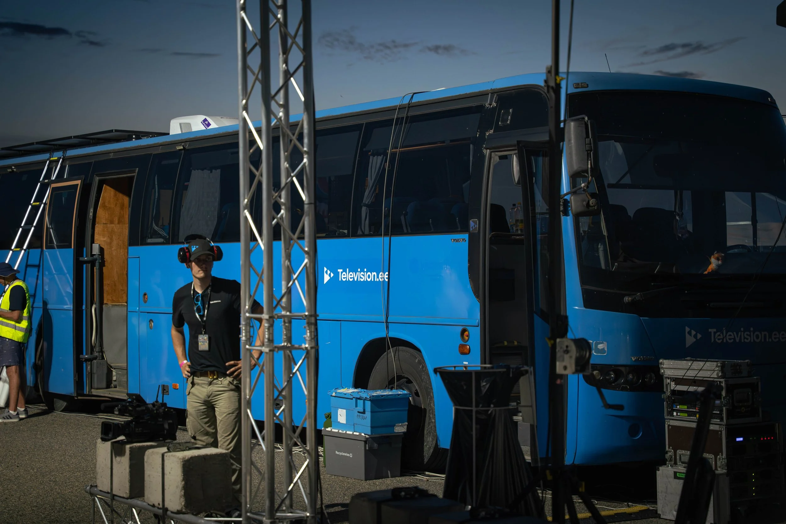 Blue broadcast bus with televisioon.ee logo, set up with equipment and crew preparing for filming or broadcasting, during dusk or evening.