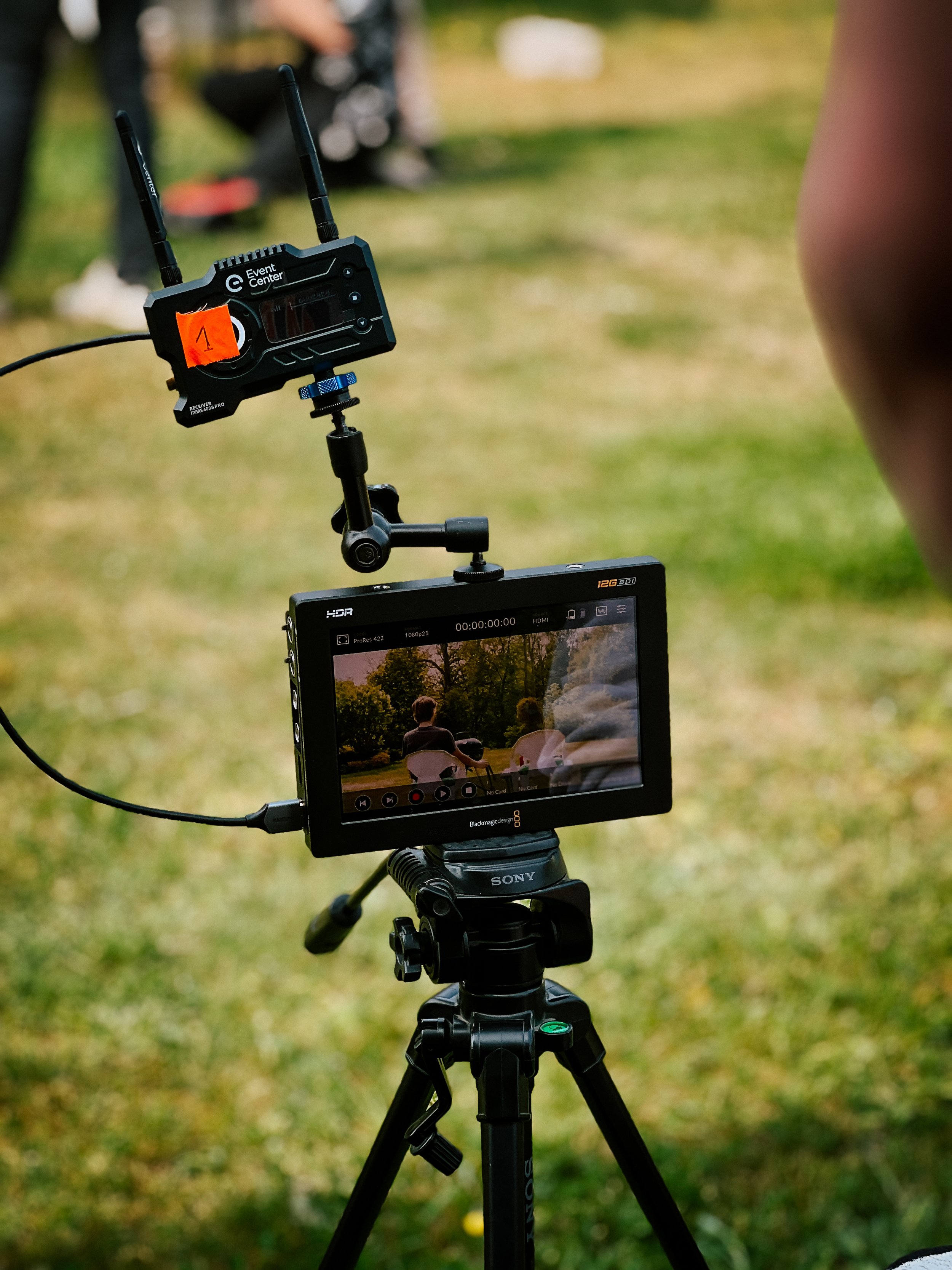 A video camera on a tripod recording two people sitting outdoors on chairs in a park or garden with trees.
