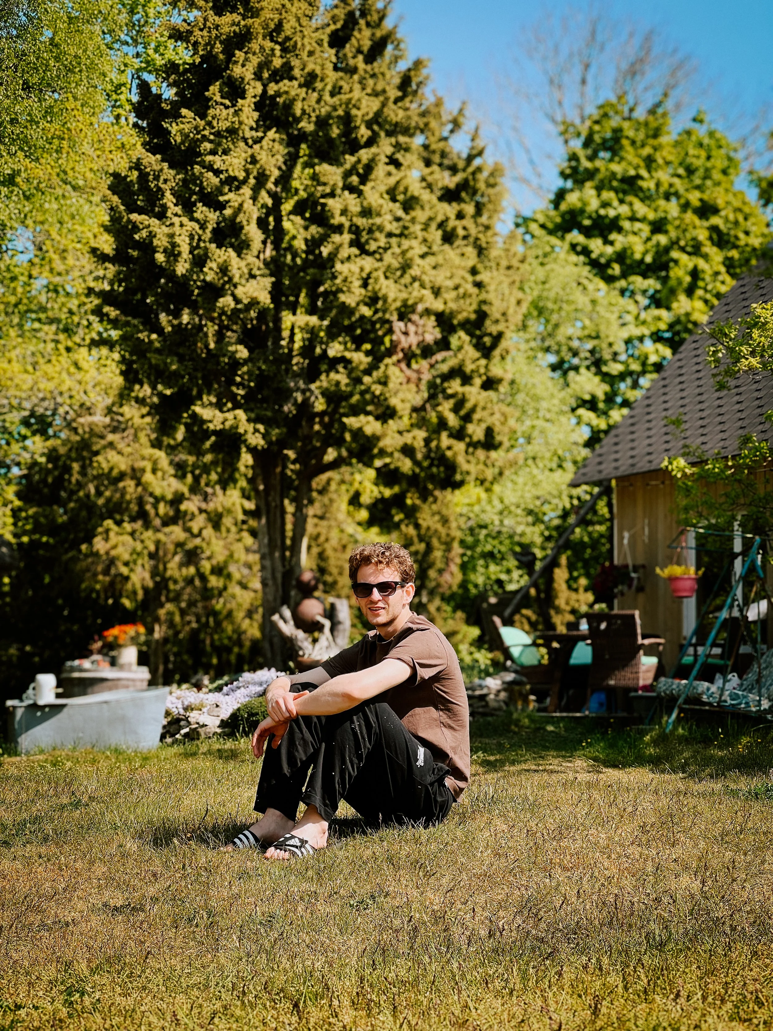 A young man with curly hair and sunglasses sitting on a grassy lawn in a backyard, with a large tree and a house with outdoor chairs and decor in the background on a sunny day.