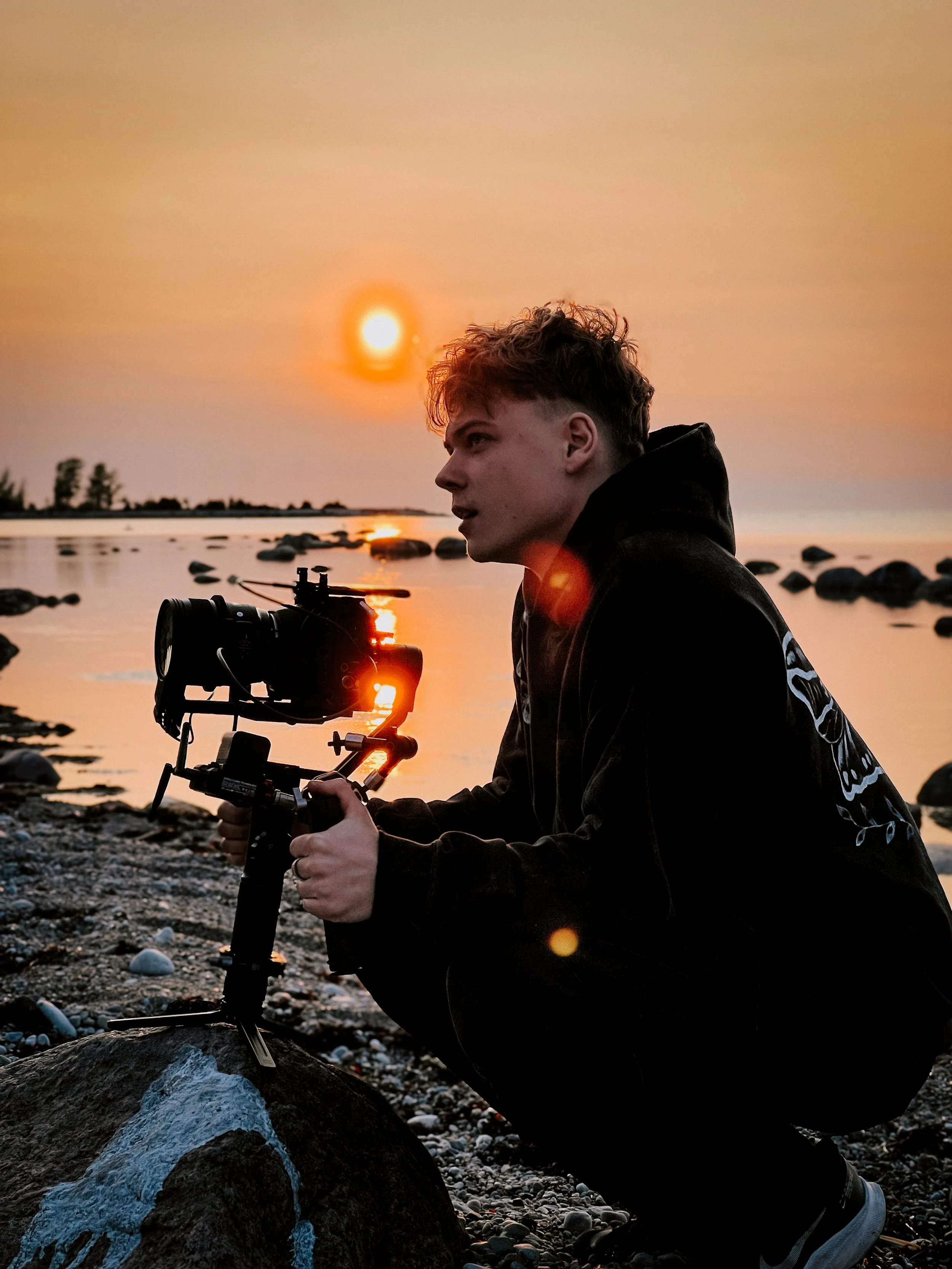 A person crouching by the water at sunset, holding a camera stabilizer, with the sun reflecting on the water and rocks on the shore.