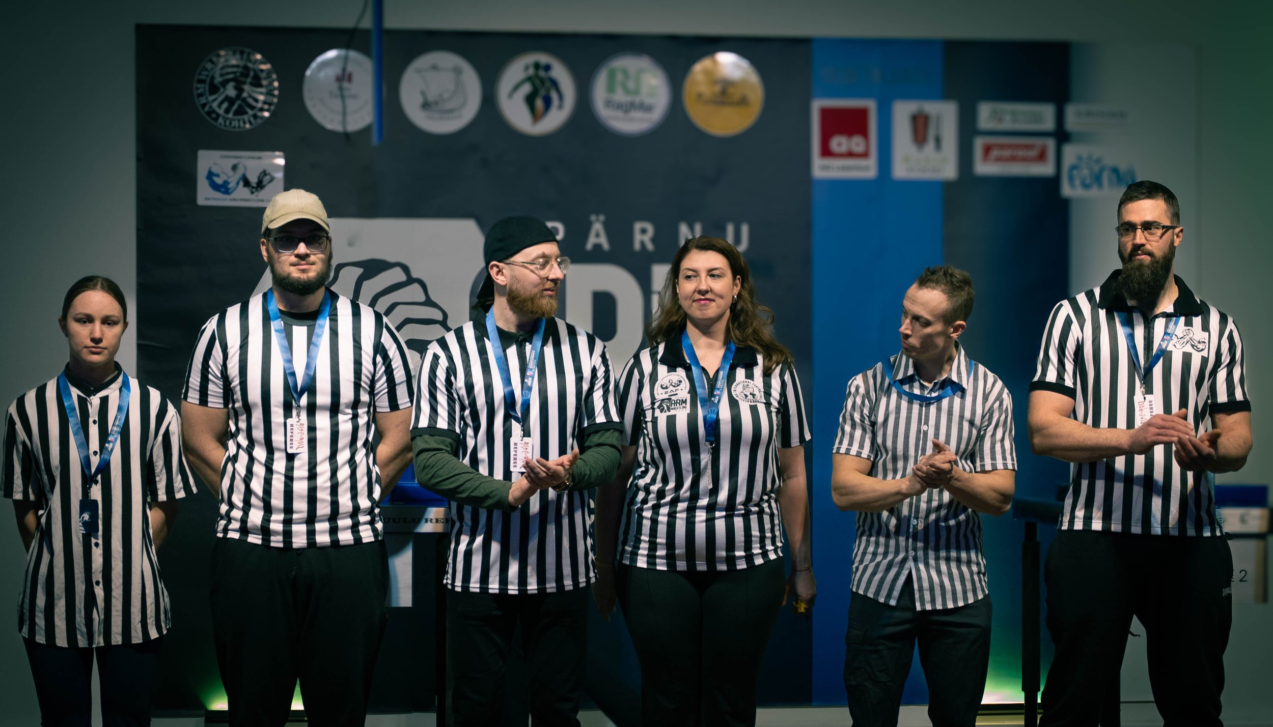 Six individuals wearing striped referee shirts stand in a line at an event, some clapping, all with blue lanyards. Behind them is a backdrop with logos and the text "PÄRNU ARM WRESTLING CLUB."