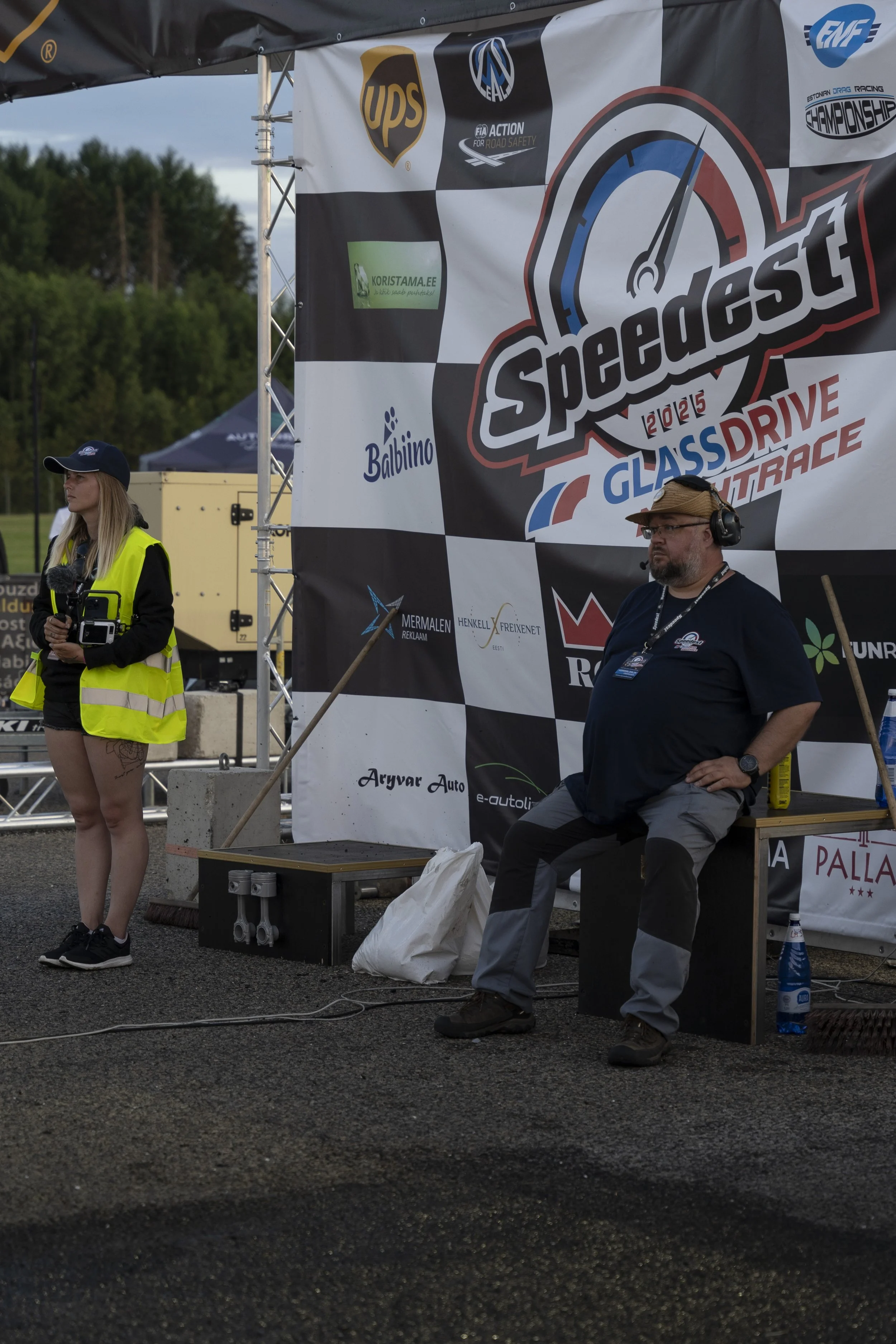 A man sitting on a bench wearing a headset, watches a race at Speedcess 2023 glass drive outdoor event, with a woman standing nearby holding a remote control, both are outdoors with greenery in the background and a banner displaying race sponsors.