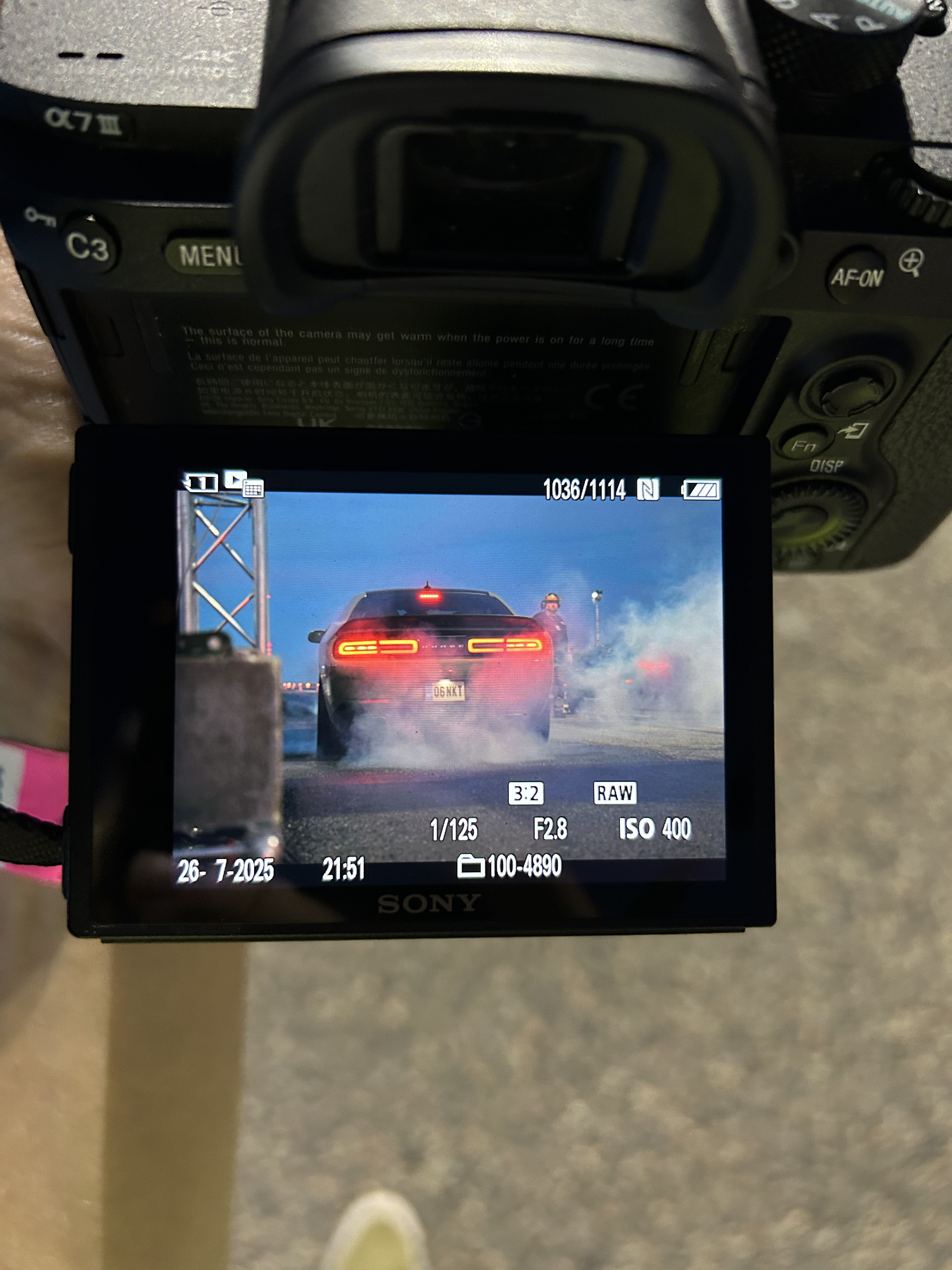 The camera screen displays a black Dodge Charger driving on a road with red brake lights illuminated, emitting smoke from the tires, with a person standing nearby in the background.
