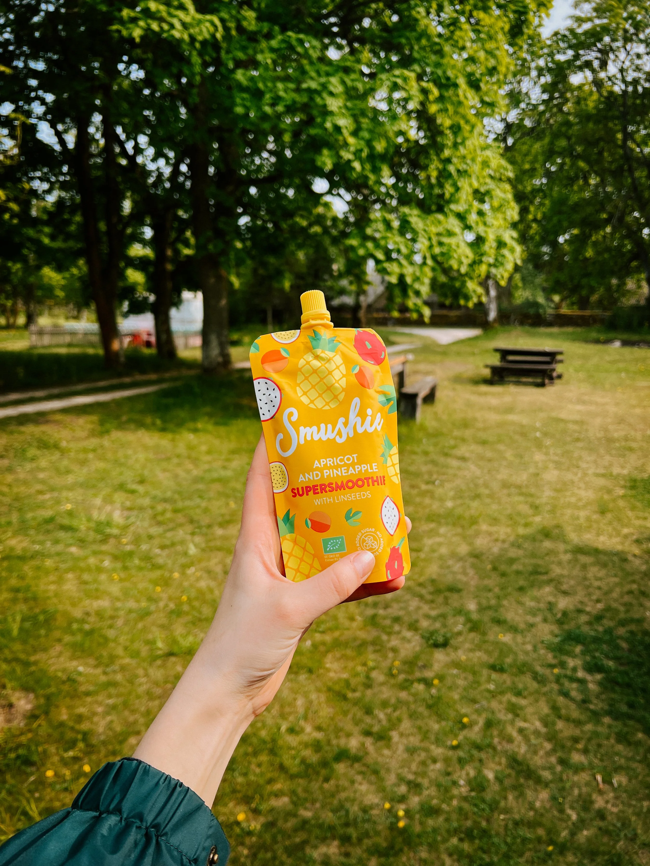 A hand holding a yellow, pineapple-shaped pouch of Smushil Apricot and Pineapple Super Smoothie with linseeds against a park with green grass, trees, and benches.