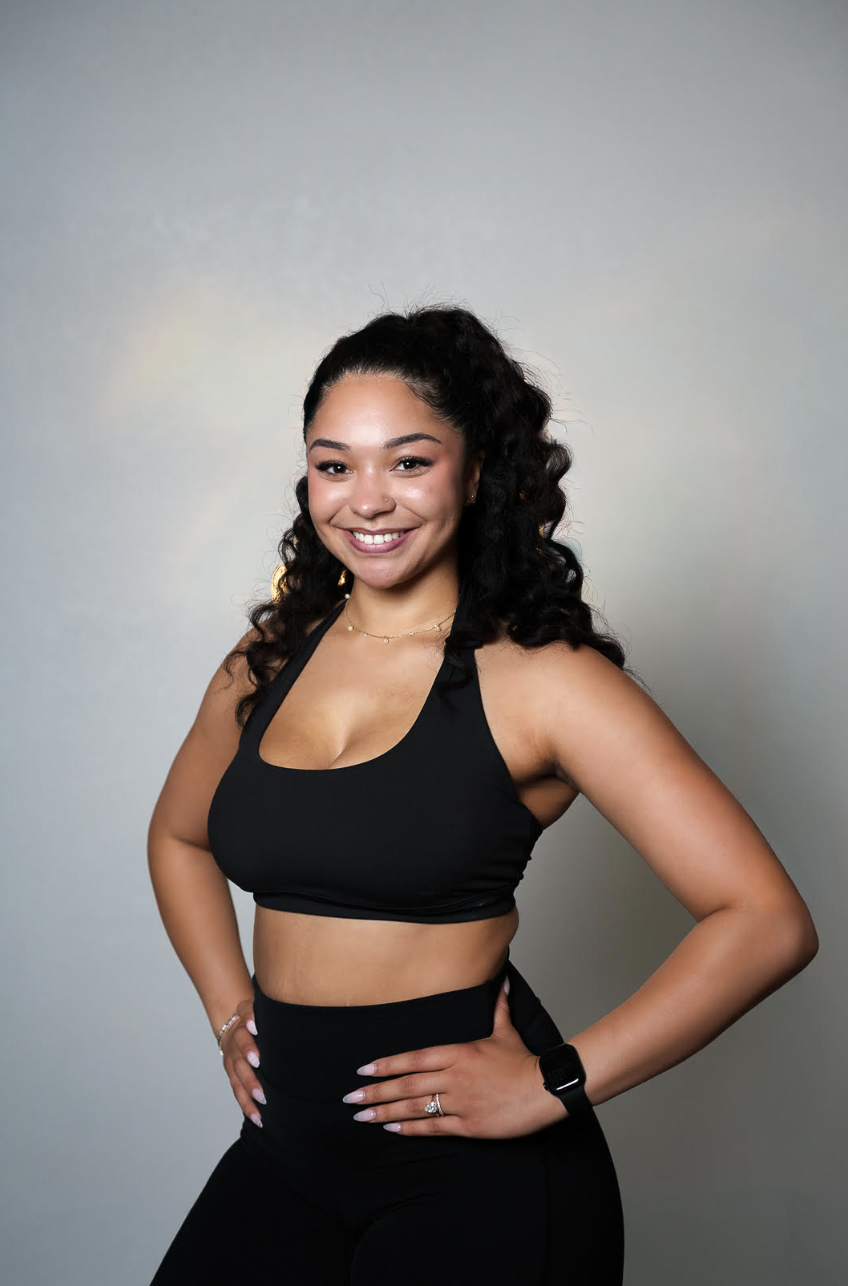 A young woman with curly dark hair wearing a black sleeveless sports top and black high-waisted leggings, smiling confidently with her hands on her hips, standing against a neutral background.