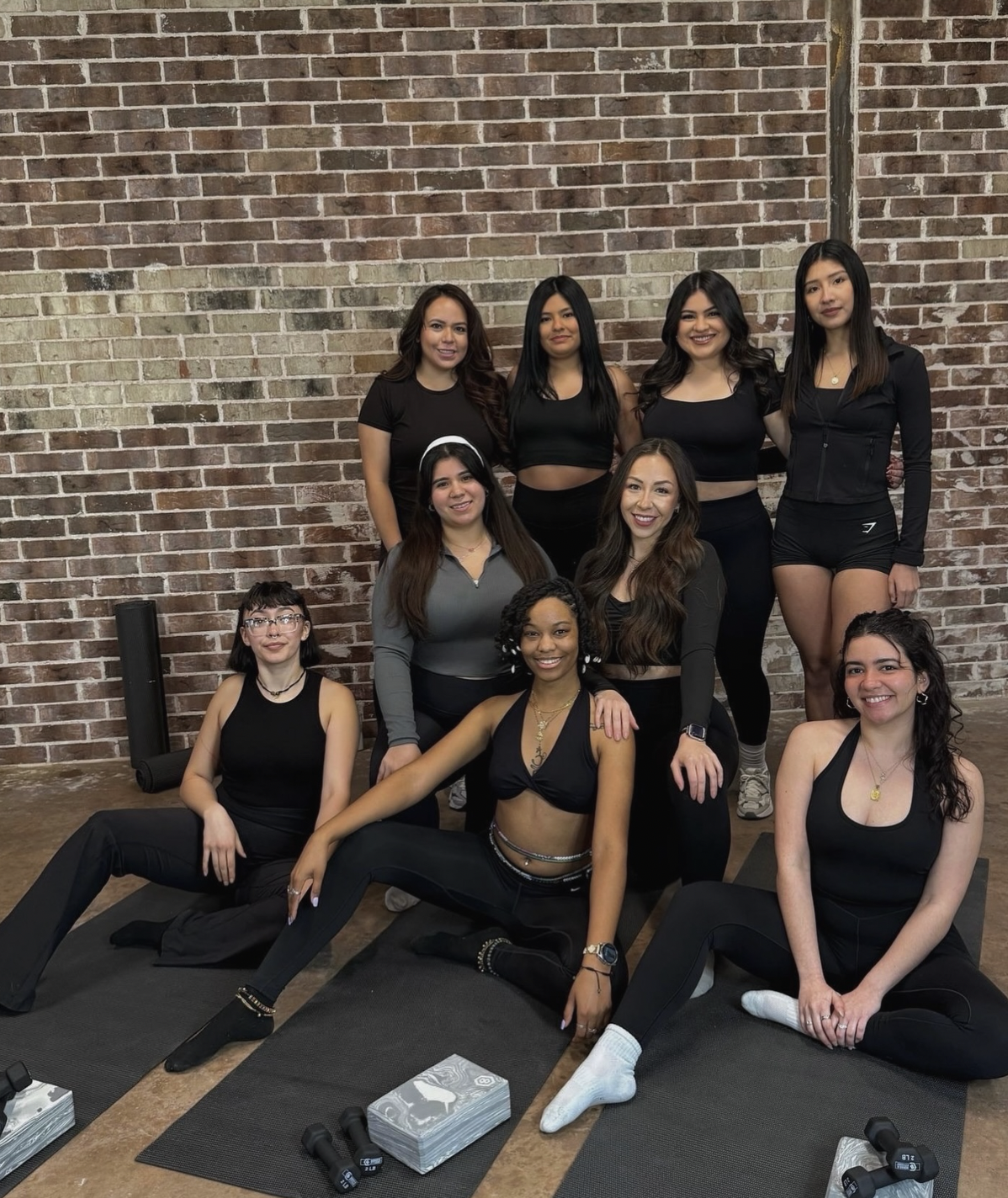 Group of women in fitness attire posing in a gym with brick wall background, some sitting on mats and others standing behind, with workout equipment and tissues in front.