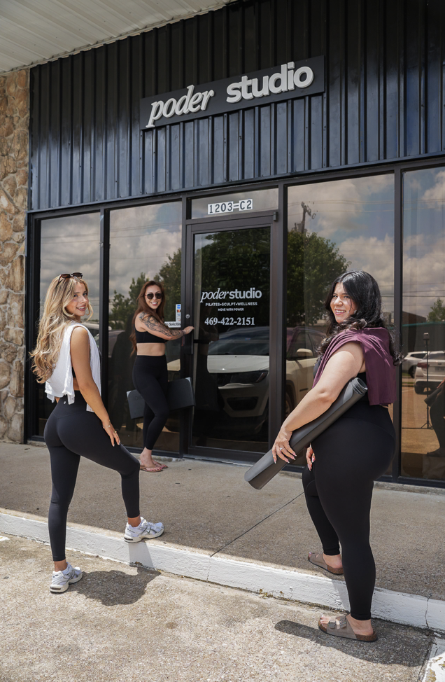 Three women standing in front of a Pilates studio, smiling and holding yoga mats, with one woman opening the door. The studio sign reads 'Poder Studio'.