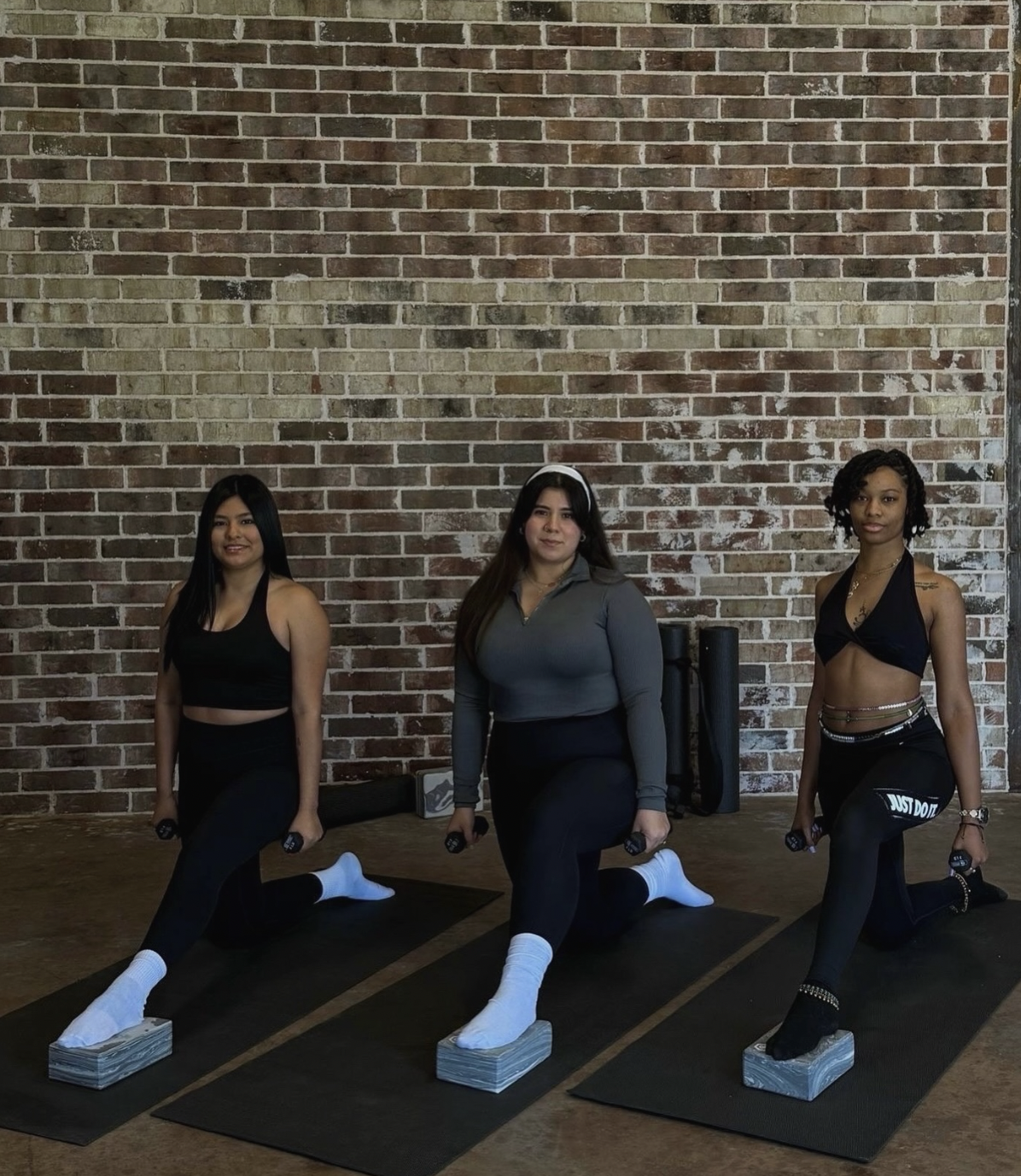 Three women kneeling on exercise mats with stacks of paper under their knees, holding weights, in a fitness studio with an exposed brick wall in the background.