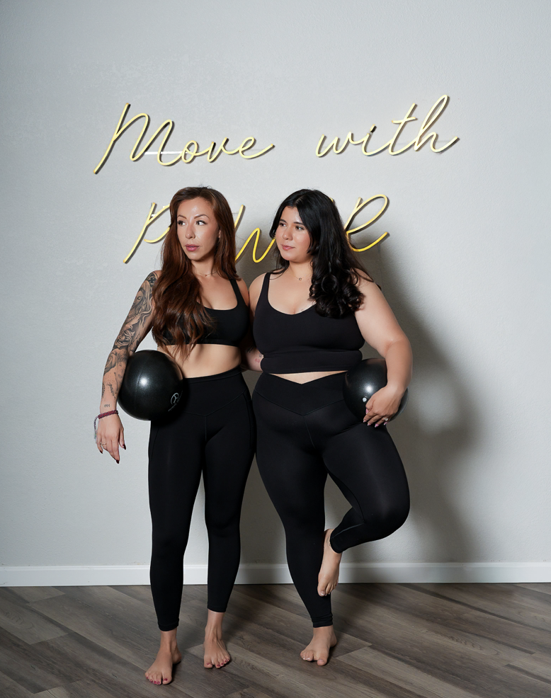 Two women in black workout clothes posing with black exercise balls in front of a wall with a gold neon sign that reads "move with"