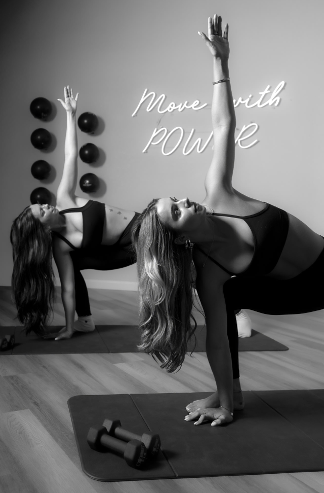 Two women practicing yoga in a studio with the phrase 'Move with Power' written on the wall. They are performing side plank poses on mats, with one of them in the foreground and the other in the background. There are dumbbells on the floor and wall-m