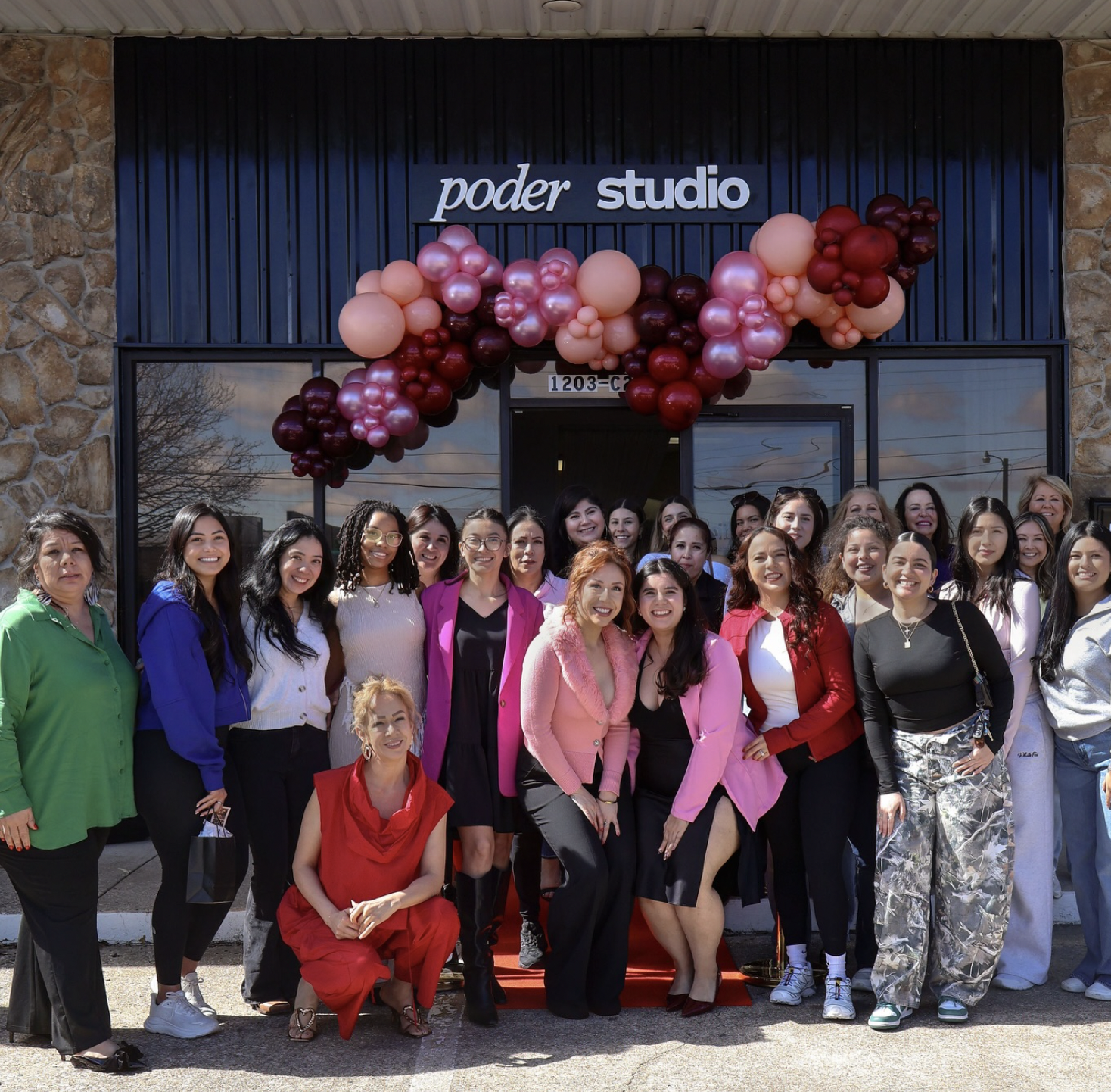 Group of women standing outside a building with a sign that reads 'poder studio,' decorated with pink and burgundy balloons, posing for a photo.