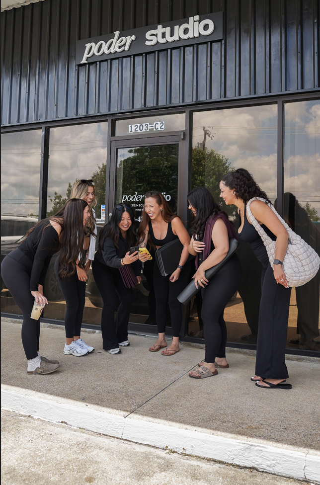 Six women standing outside a building with a sign that reads "poder studio." They are gathered around a smartphone, smiling and looking at the screen, appearing to share a joyful moment.
