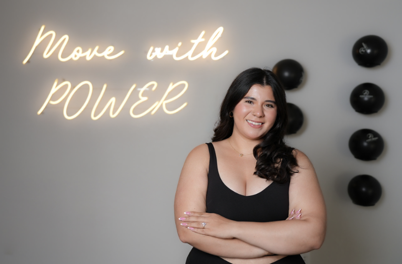 A woman standing with arms crossed in front of a gray wall with a neon sign that says 'Move with POWER' and black exercise balls to her right.
