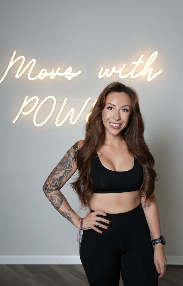 A smiling woman in athletic wear standing in front of a wall with neon sign that says 'Move with Power'.