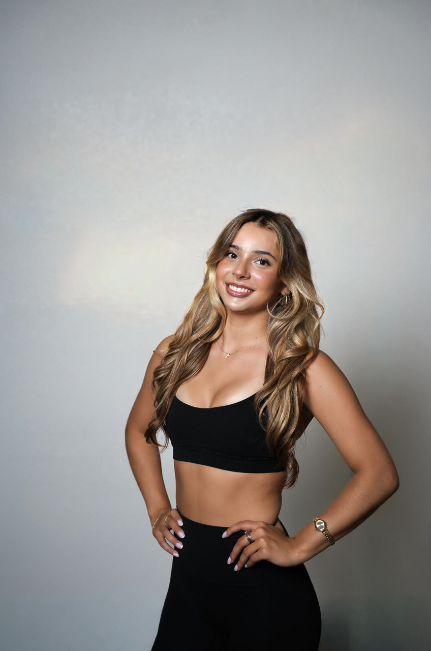 A young woman with long wavy blonde hair, wearing a black sports bra and black high-waisted pants, smiling and standing with hands on hips against a plain light gray background.