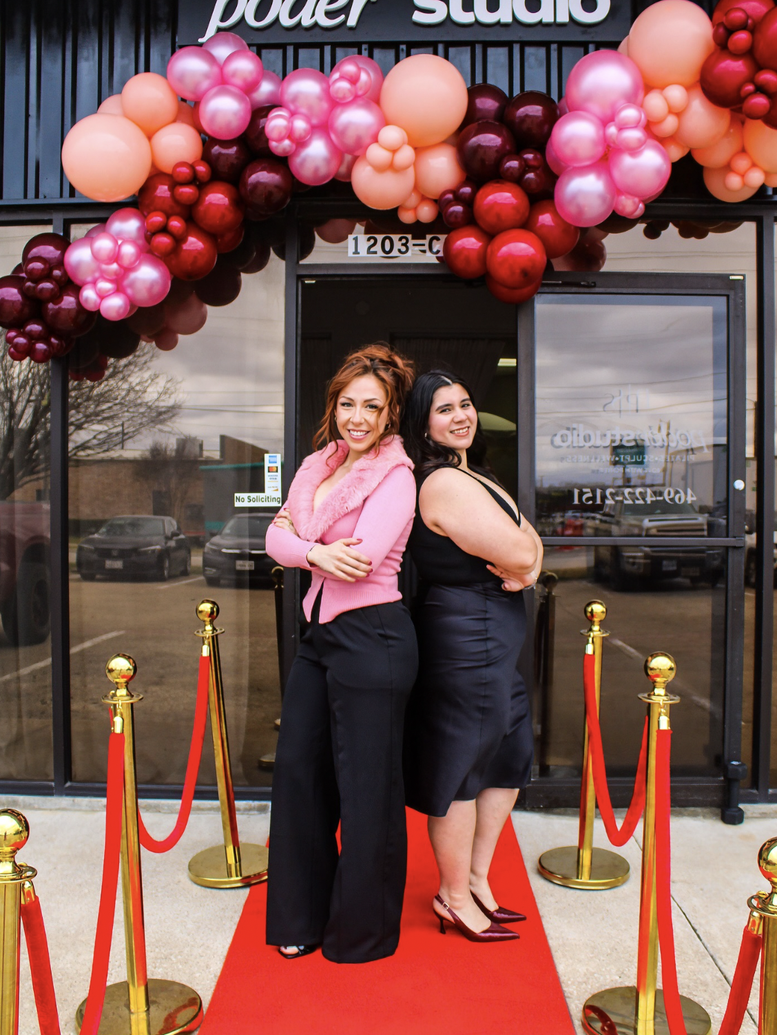 Two women standing on a red carpet in front of a store entrance decorated with pink, red, and peach balloons. The women are smiling and posing with their arms crossed.