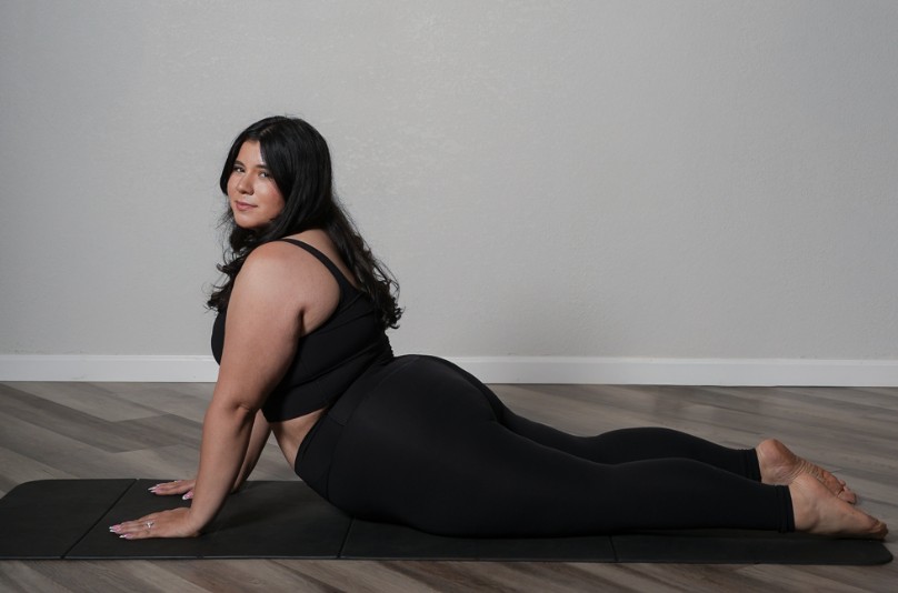 A woman performing a yoga pose on a black mat, stretching on a hardwood floor in a plain room.