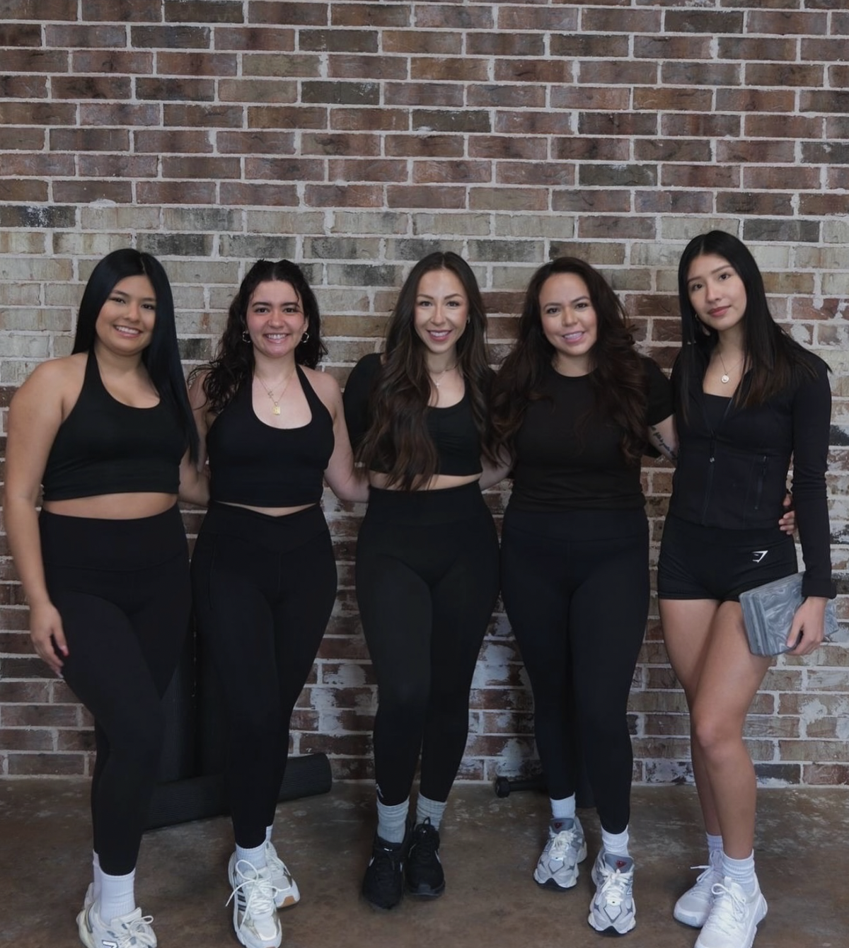 Five women standing in front of a brick wall, dressed in workout clothes, smiling at the camera.