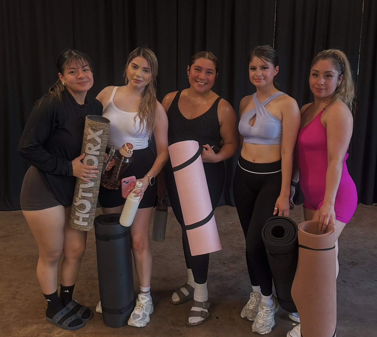 Group of five women in athletic wear holding yoga mats and water bottles, standing on a wooden floor in front of a dark curtain, smiling after a fitness class.