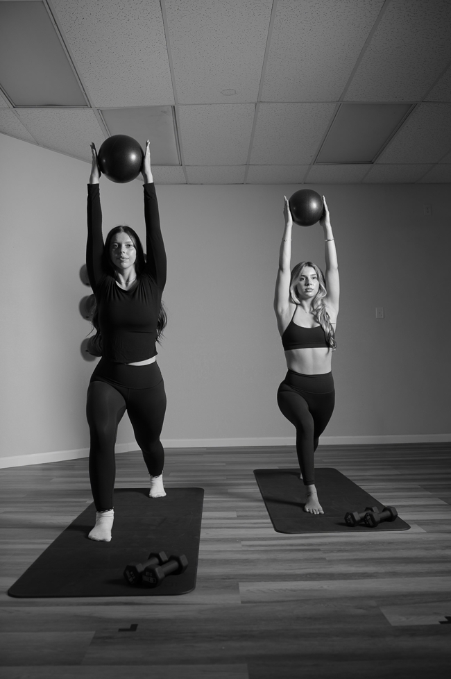 Two women in workout clothes standing on exercise mats, lifting medicine balls overhead in a fitness studio with wooden floors and a ceiling with acoustic tiles.